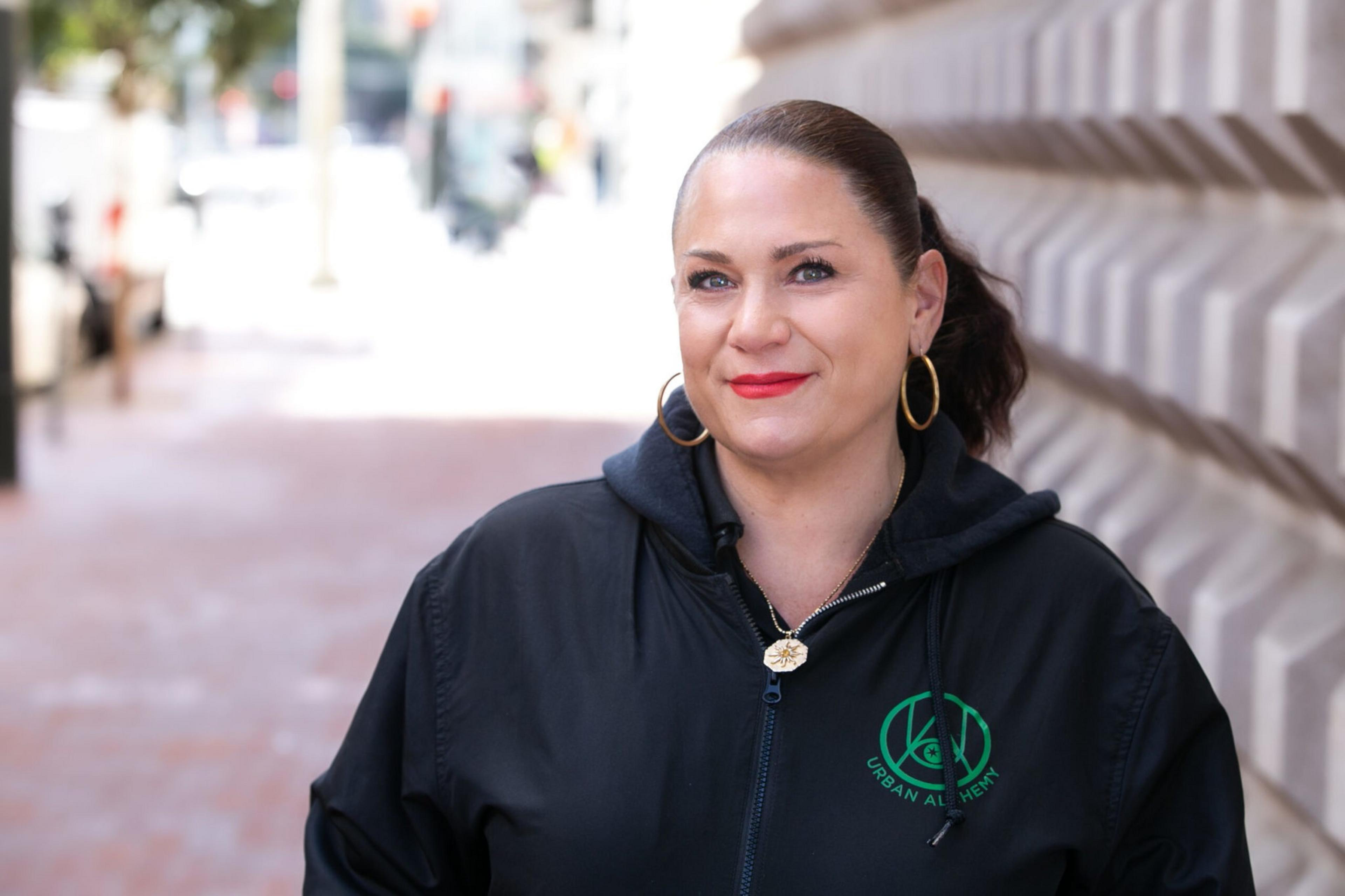 A woman with slicked-back hair, red lipstick, and gold hoop earrings smiles while wearing a black Urban Alchemy jacket, standing outdoors near a textured wall.