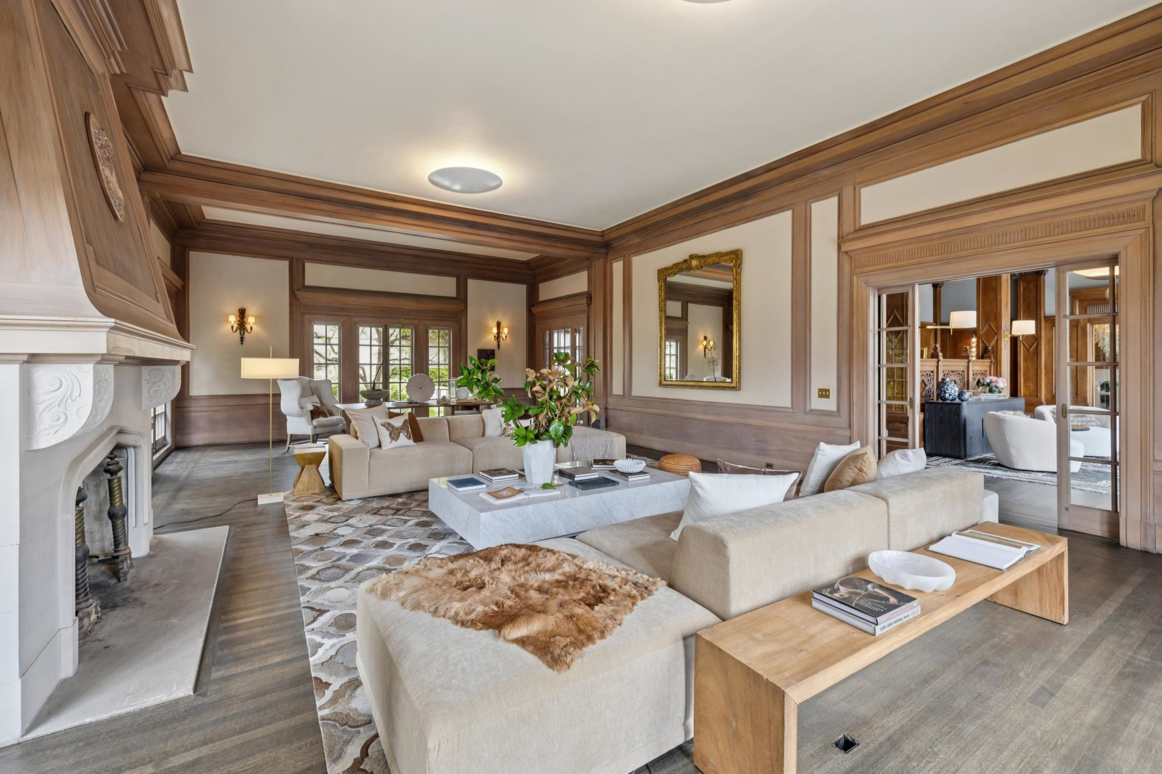 A spacious living room features beige sofas, a stone fireplace, a large mirror, wooden trim, a geometric rug, and various decorative plants and books.
