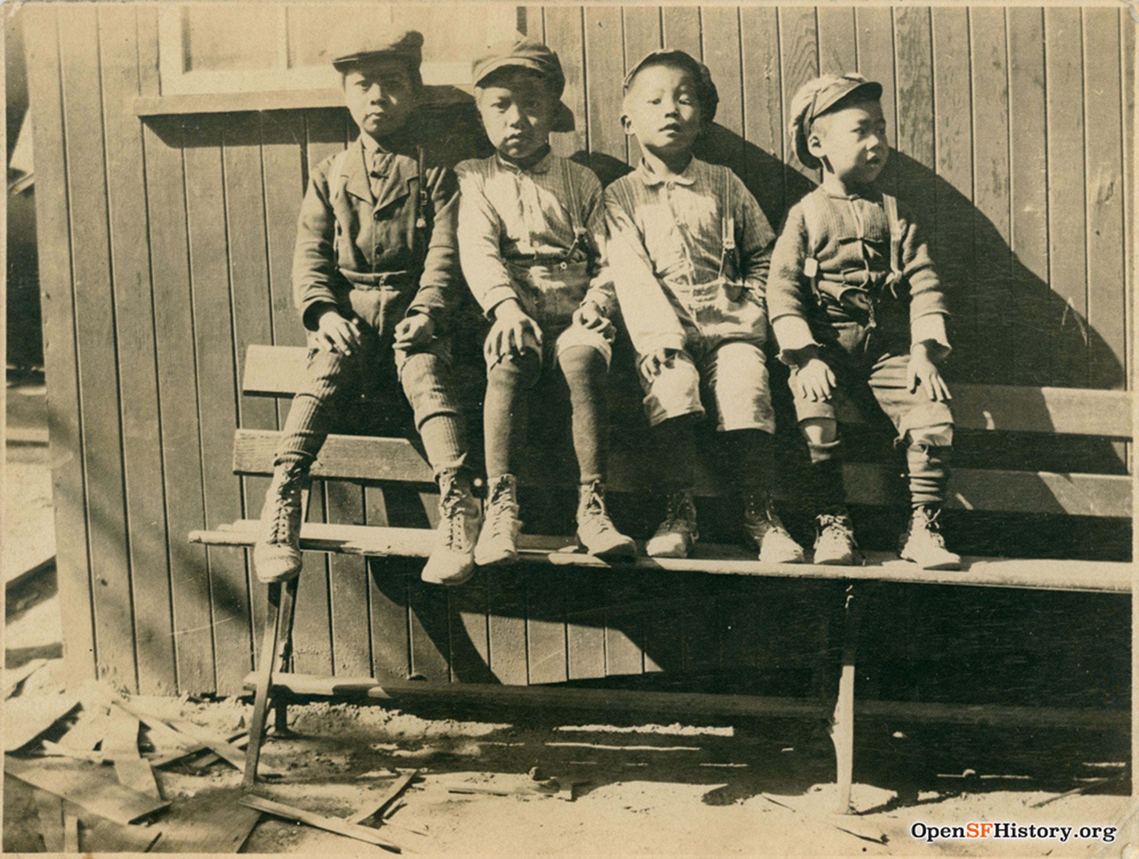 Four young boys wearing old-fashioned clothes and caps sit side by side on a wooden bench against a wall, with shadows cast behind them.