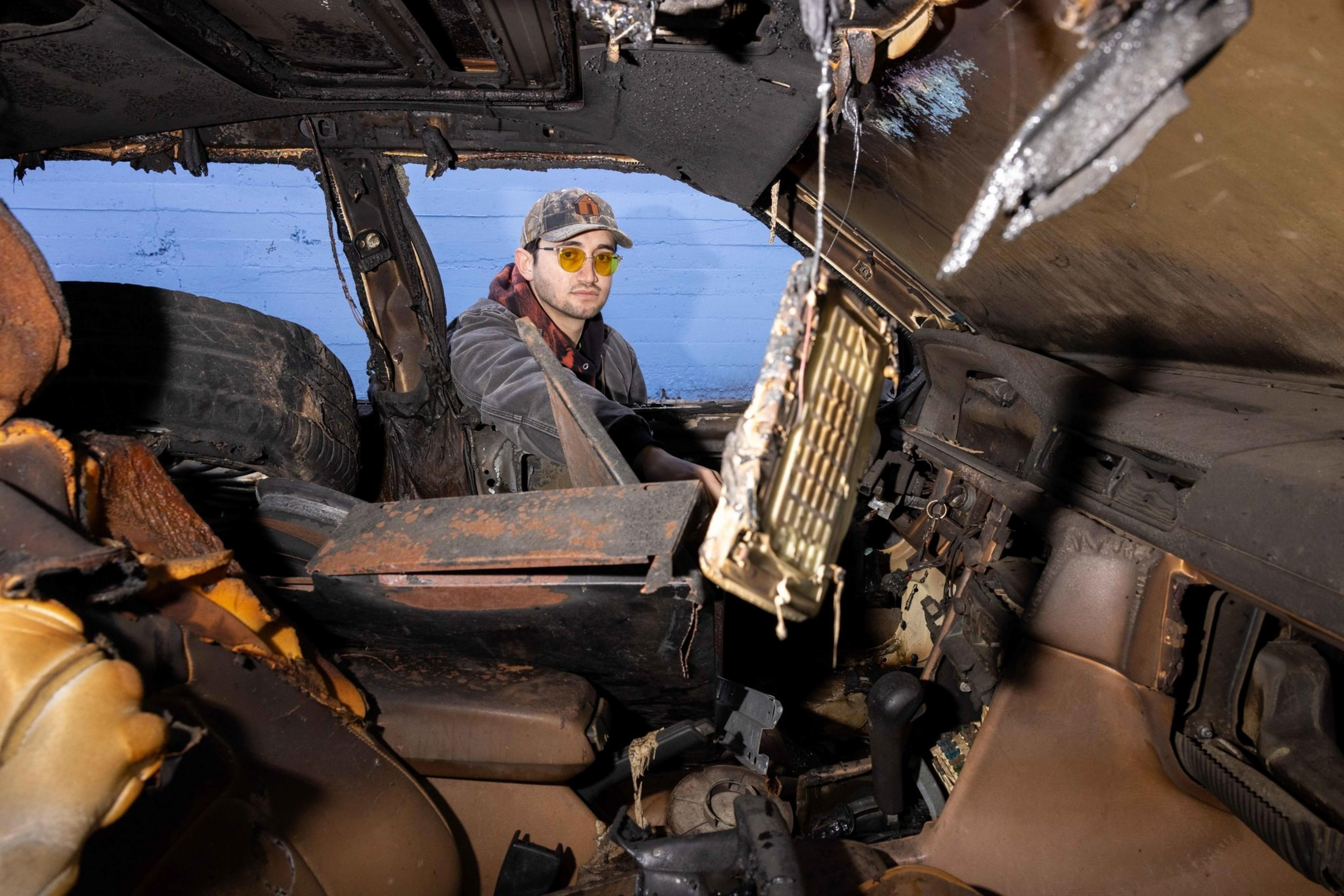 A person wearing a cap and yellow glasses sits in a severely damaged and burnt car interior, with exposed metal and debris around them.