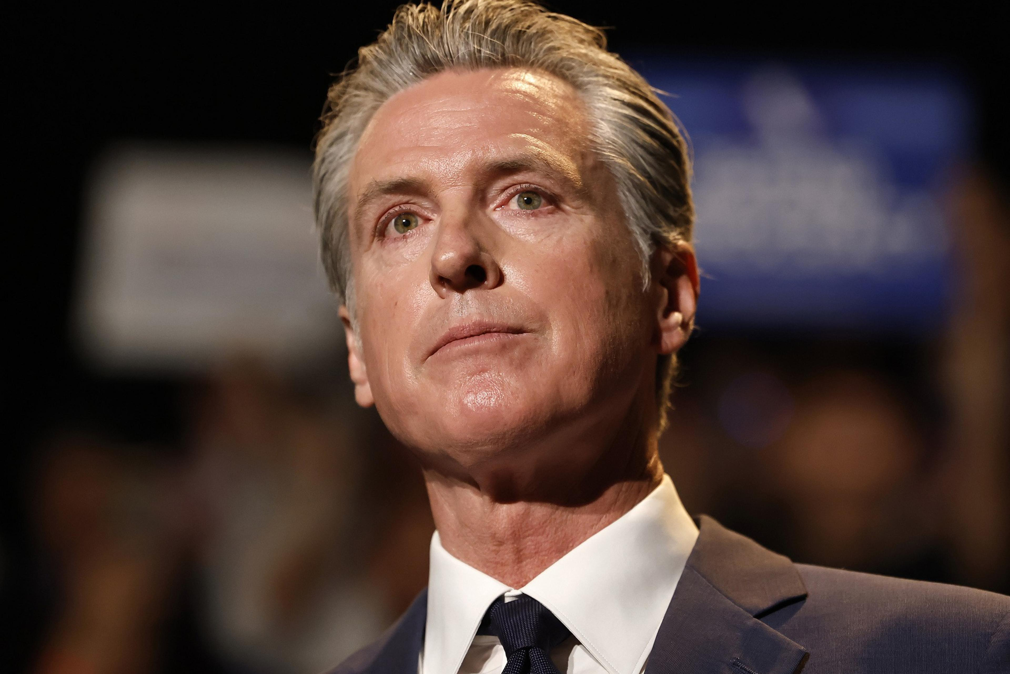 A middle-aged man with slicked-back gray hair wears a dark suit and tie, looking thoughtfully upward against a blurred background.