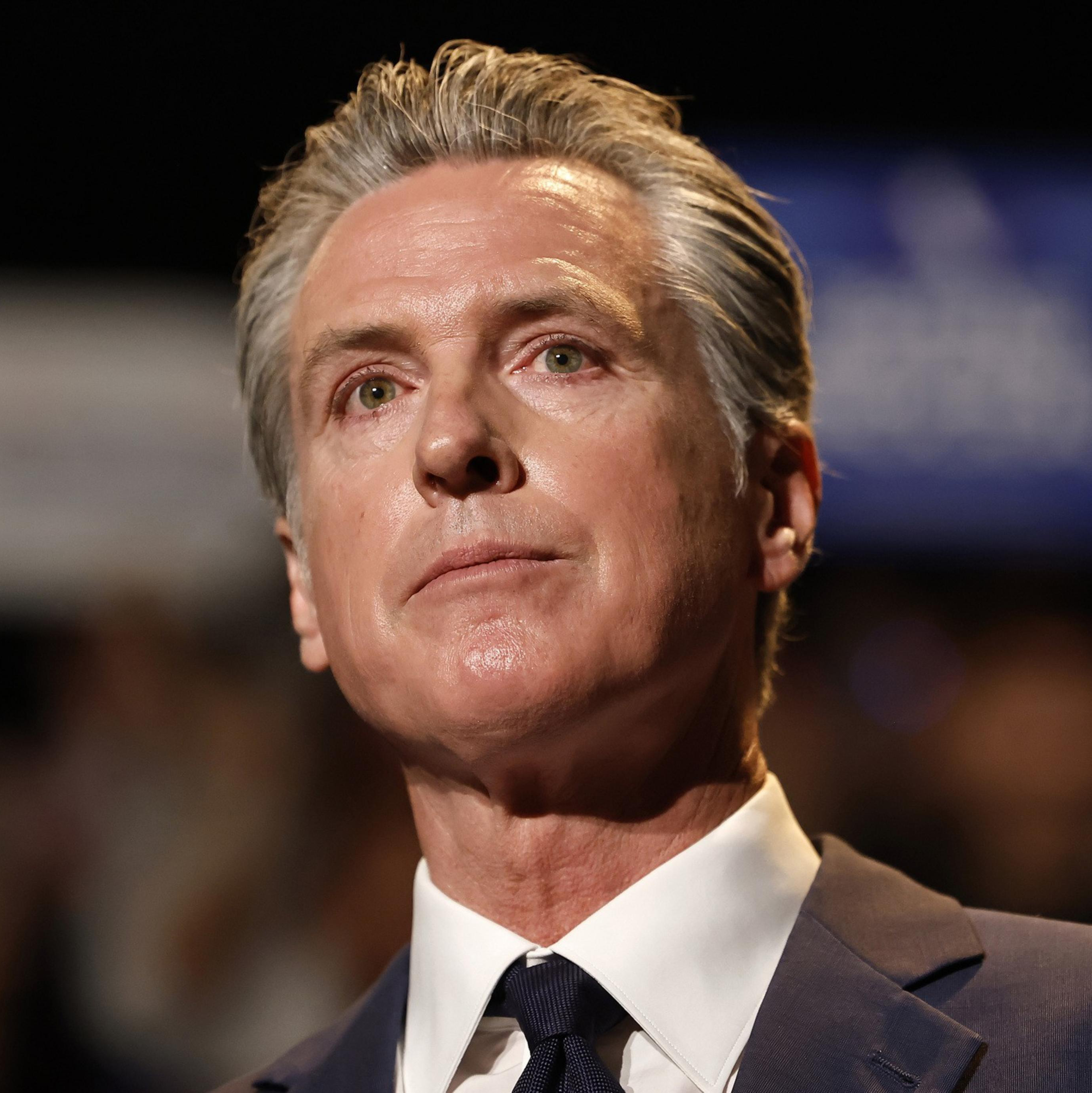 A middle-aged man with slicked-back gray hair wears a dark suit and tie, looking thoughtfully upward against a blurred background.