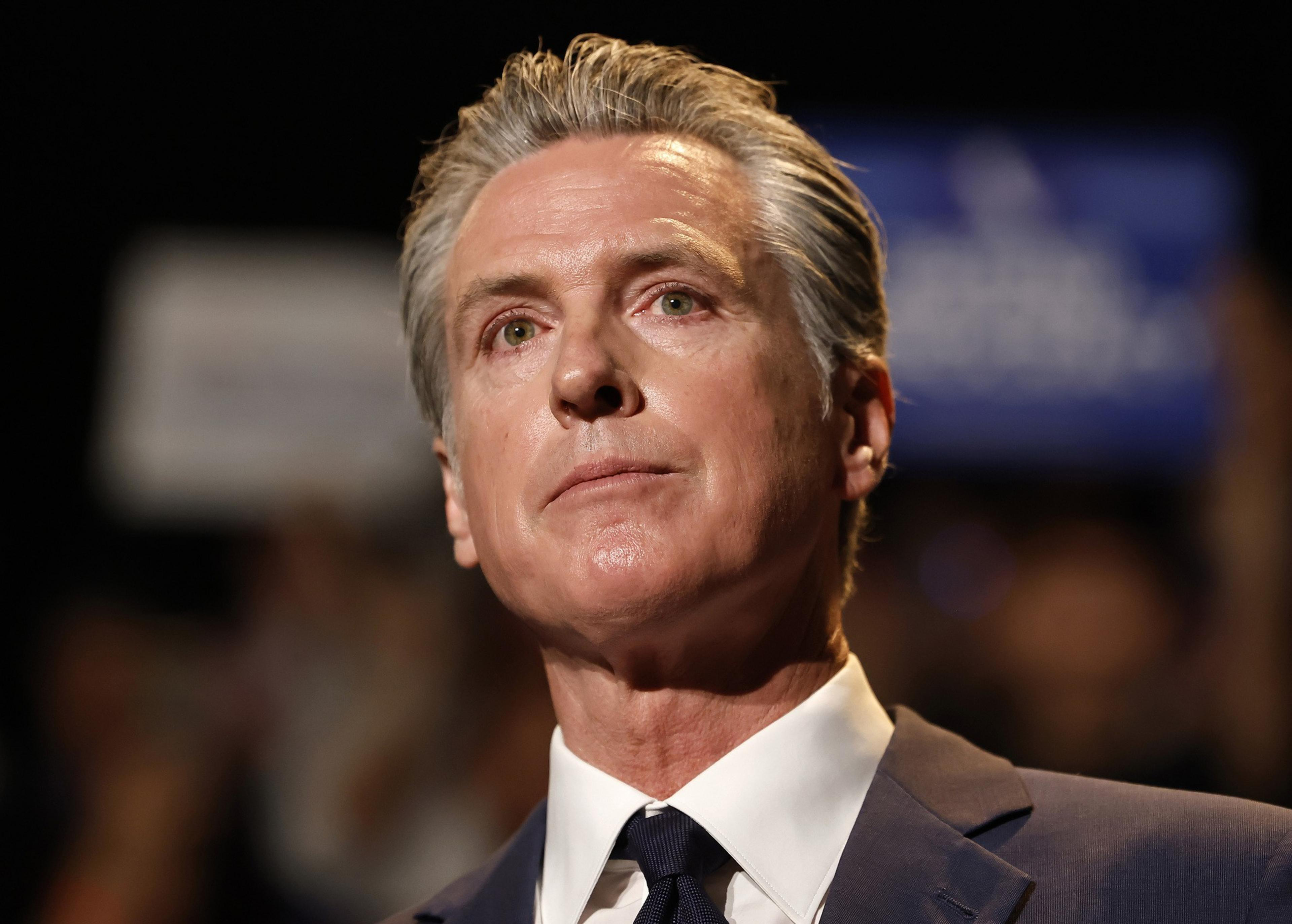 A middle-aged man with slicked-back gray hair wears a dark suit and tie, looking thoughtfully upward against a blurred background.