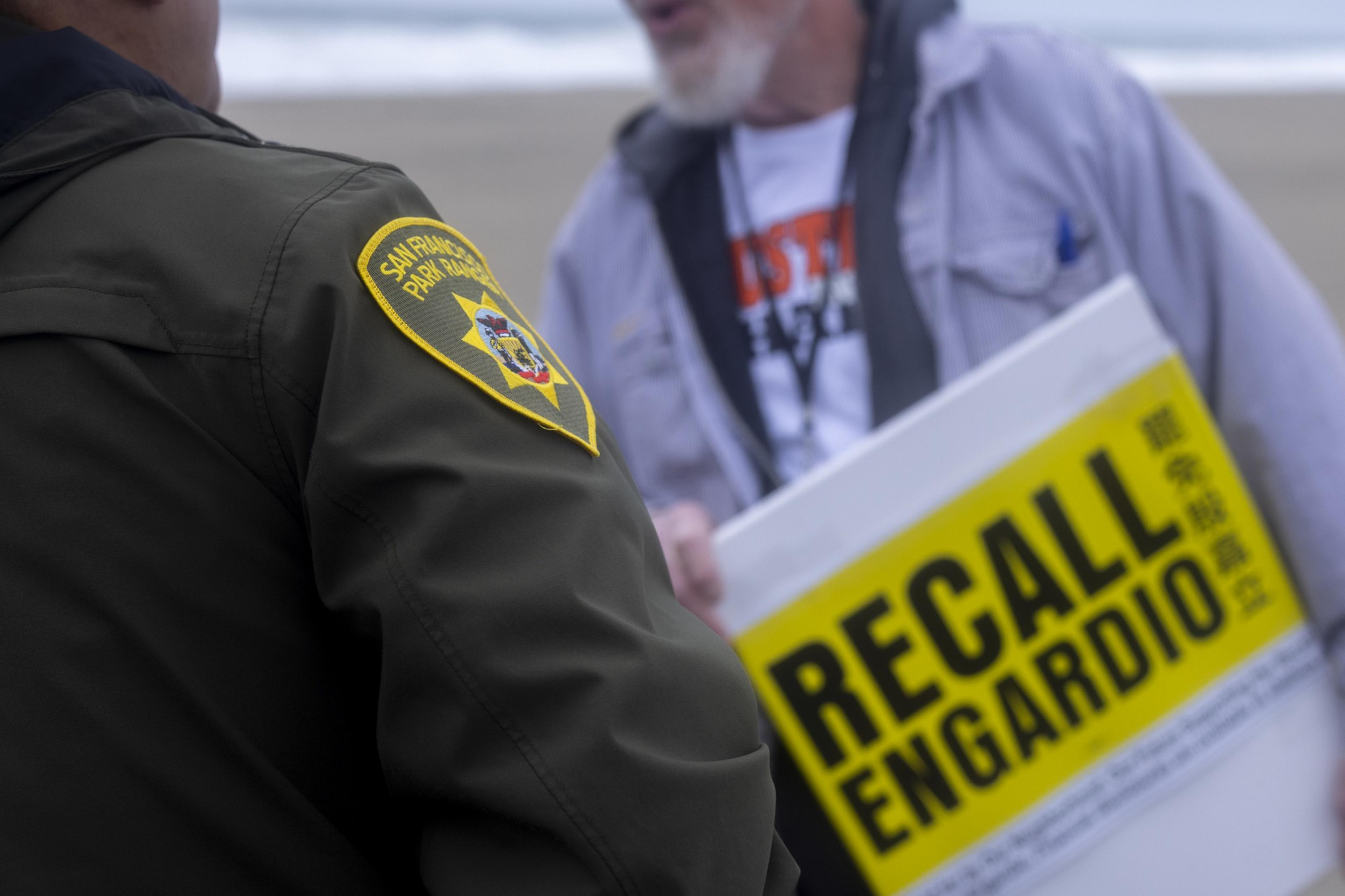 A person wearing a &quot;San Francisco Park Ranger&quot; jacket stands beside another holding a yellow sign that reads &quot;RECALL ENGARDIO&quot; at a beach setting.