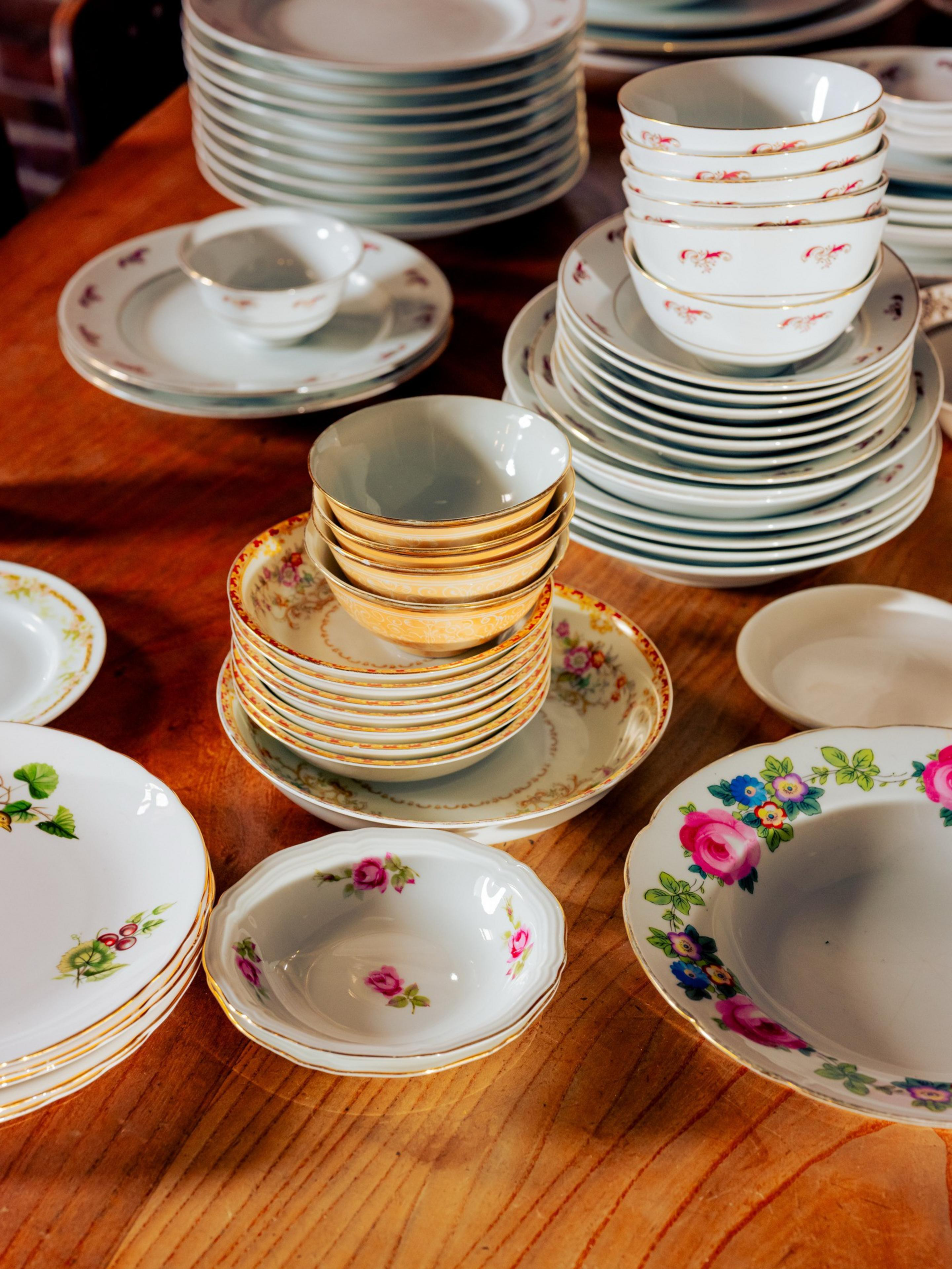 A variety of stacked porcelain dishes with floral patterns sit on a wooden table. The sets include bowls, plates, and small saucers, all decorated with colorful flowers.