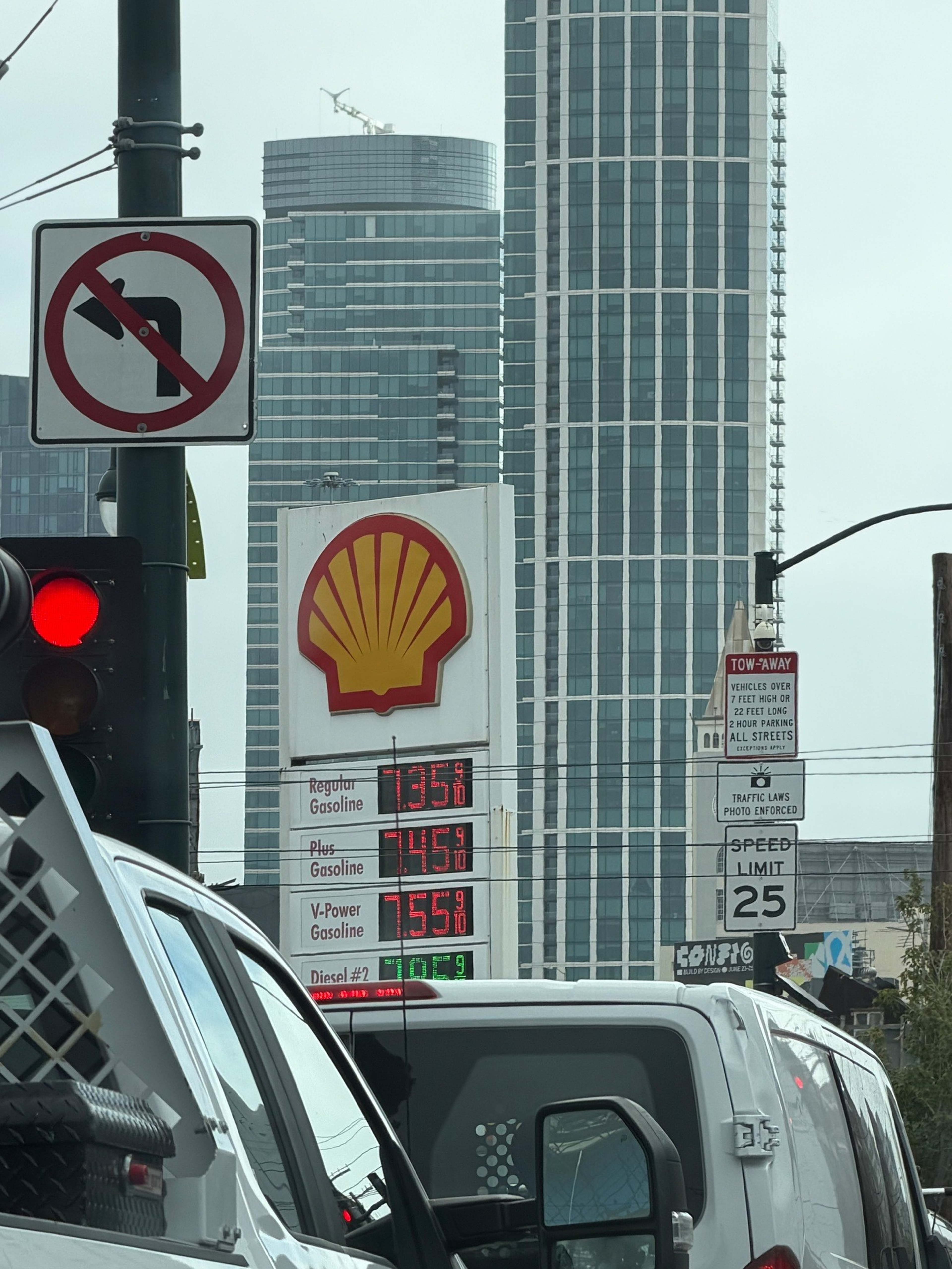 Gas prices at a Shell station read $7.35, $7.45, and $7.55 per gallon with a no left turn sign, red traffic light, and speed limit 25 sign visible.
