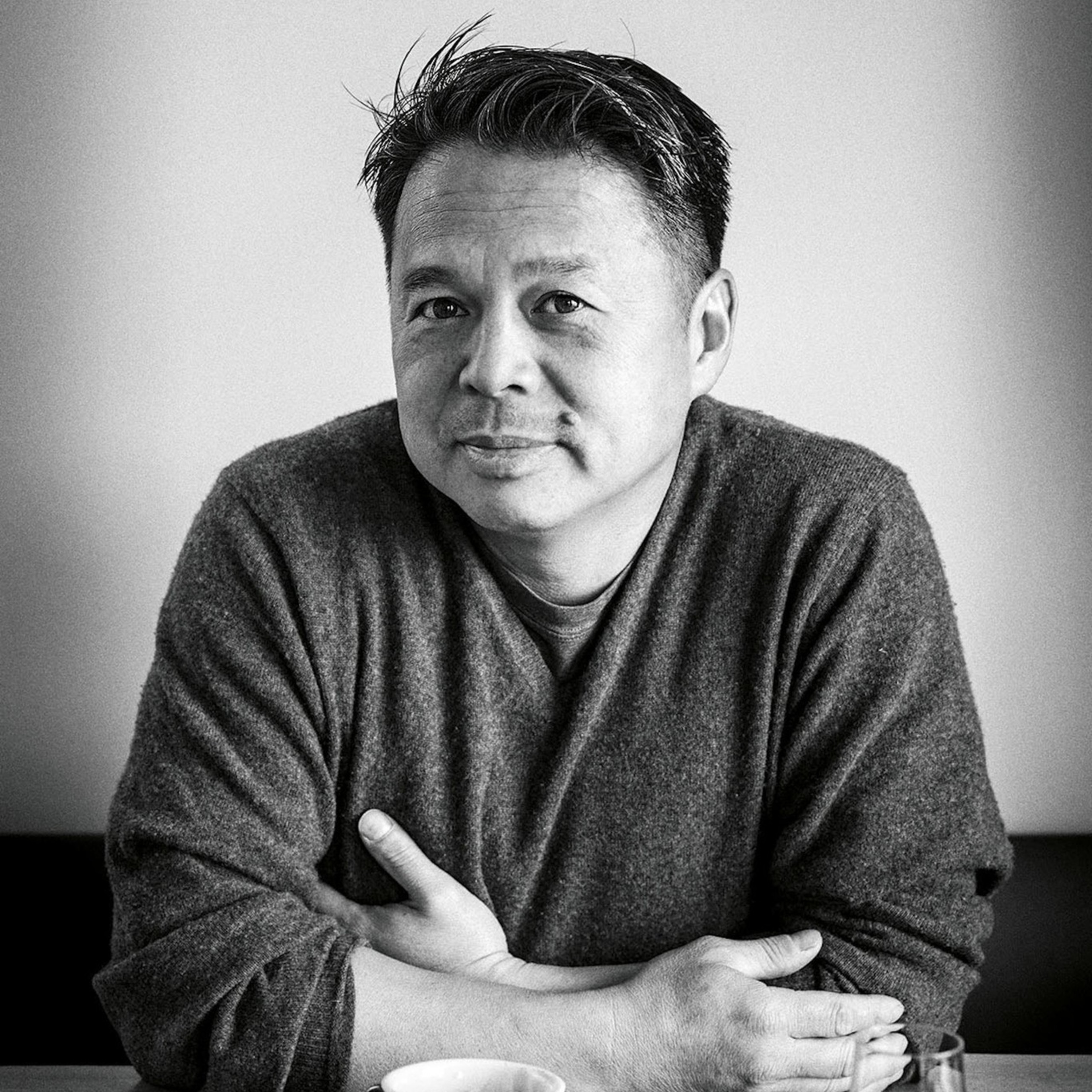 A man wearing a dark sweater sits at a table with a mug of coffee.
