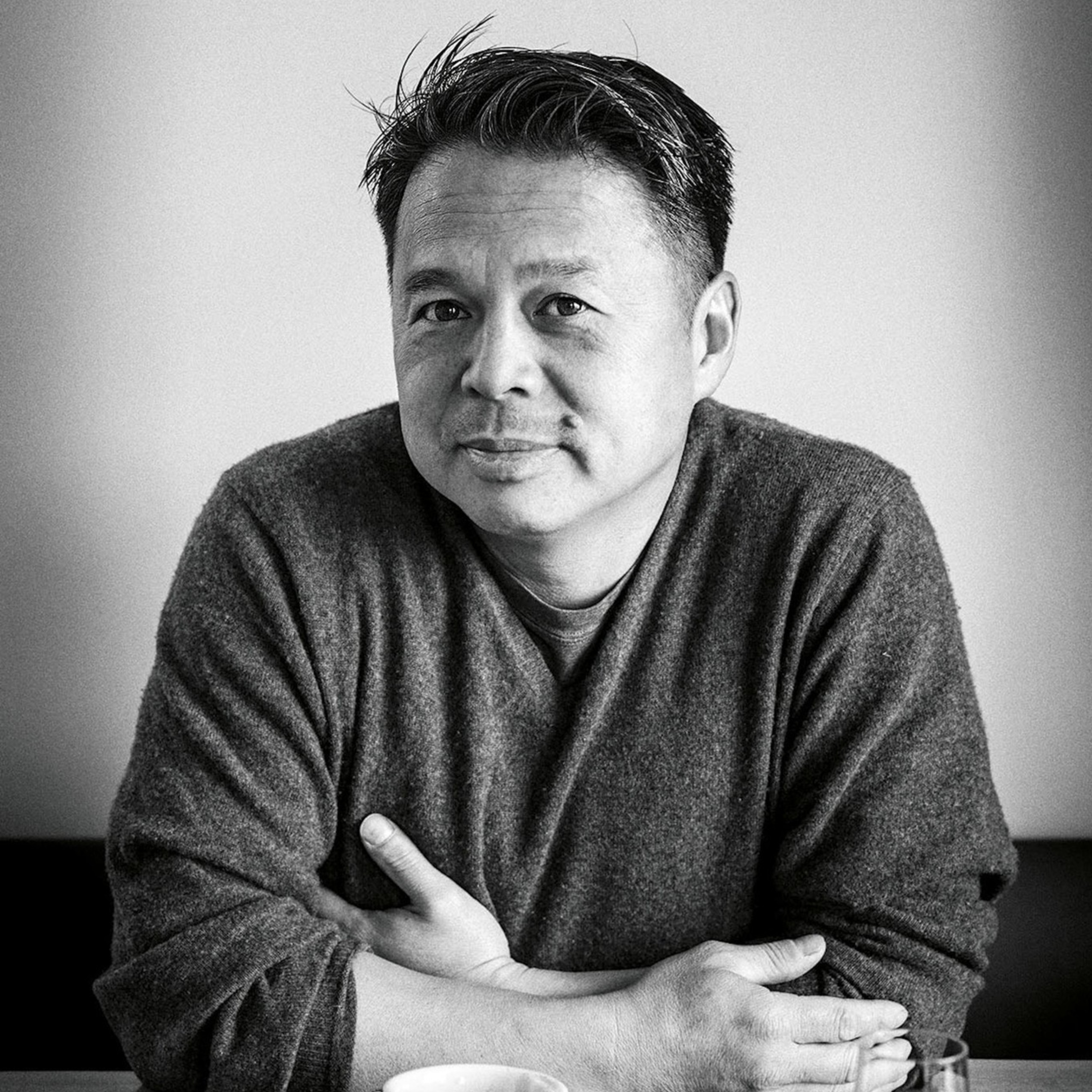 A man wearing a dark sweater sits at a table with a mug of coffee.