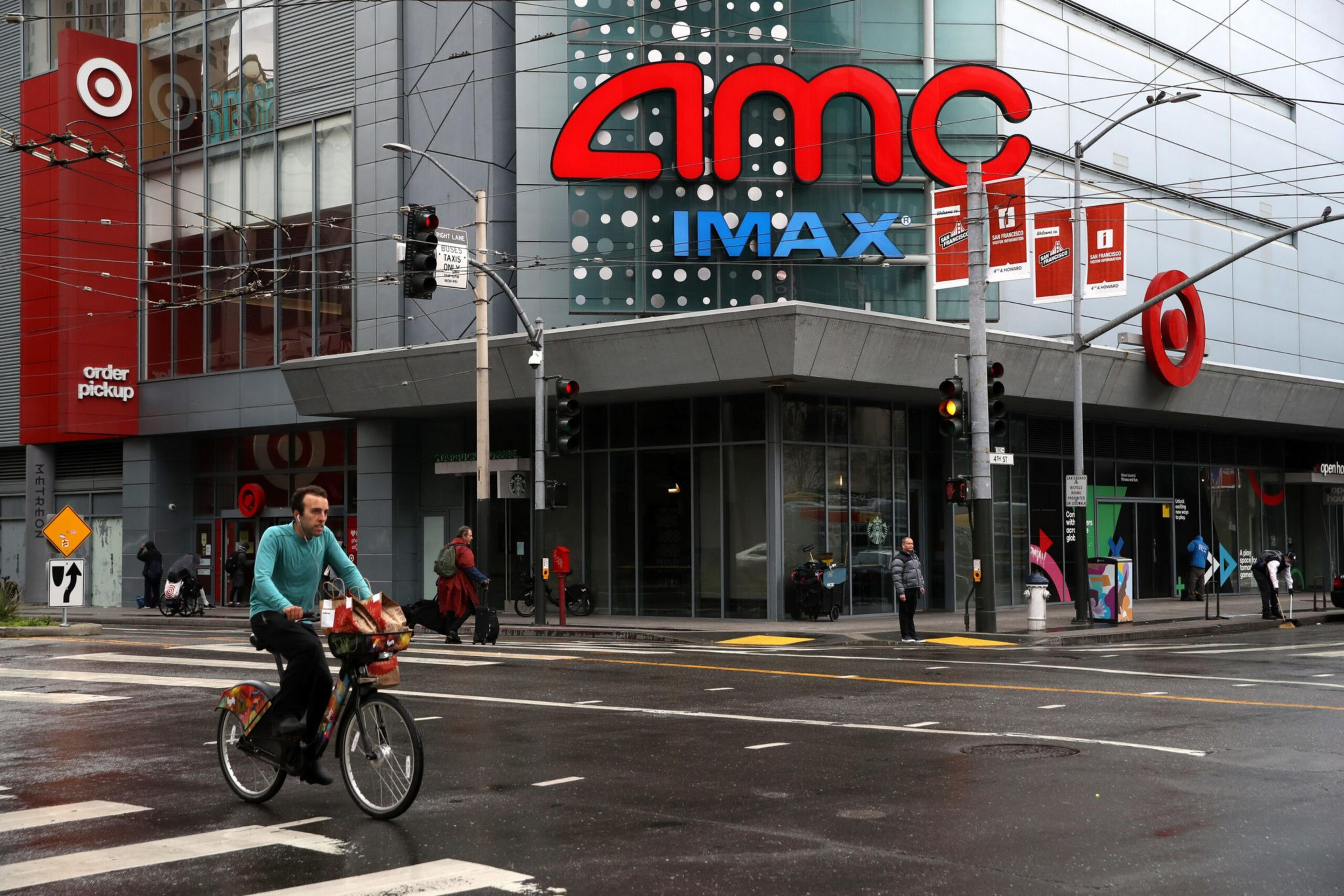 A man rides a bicycle across a wet street in front of an AMC IMAX theater and a Target store. Pedestrians walk nearby, and the traffic lights are red.