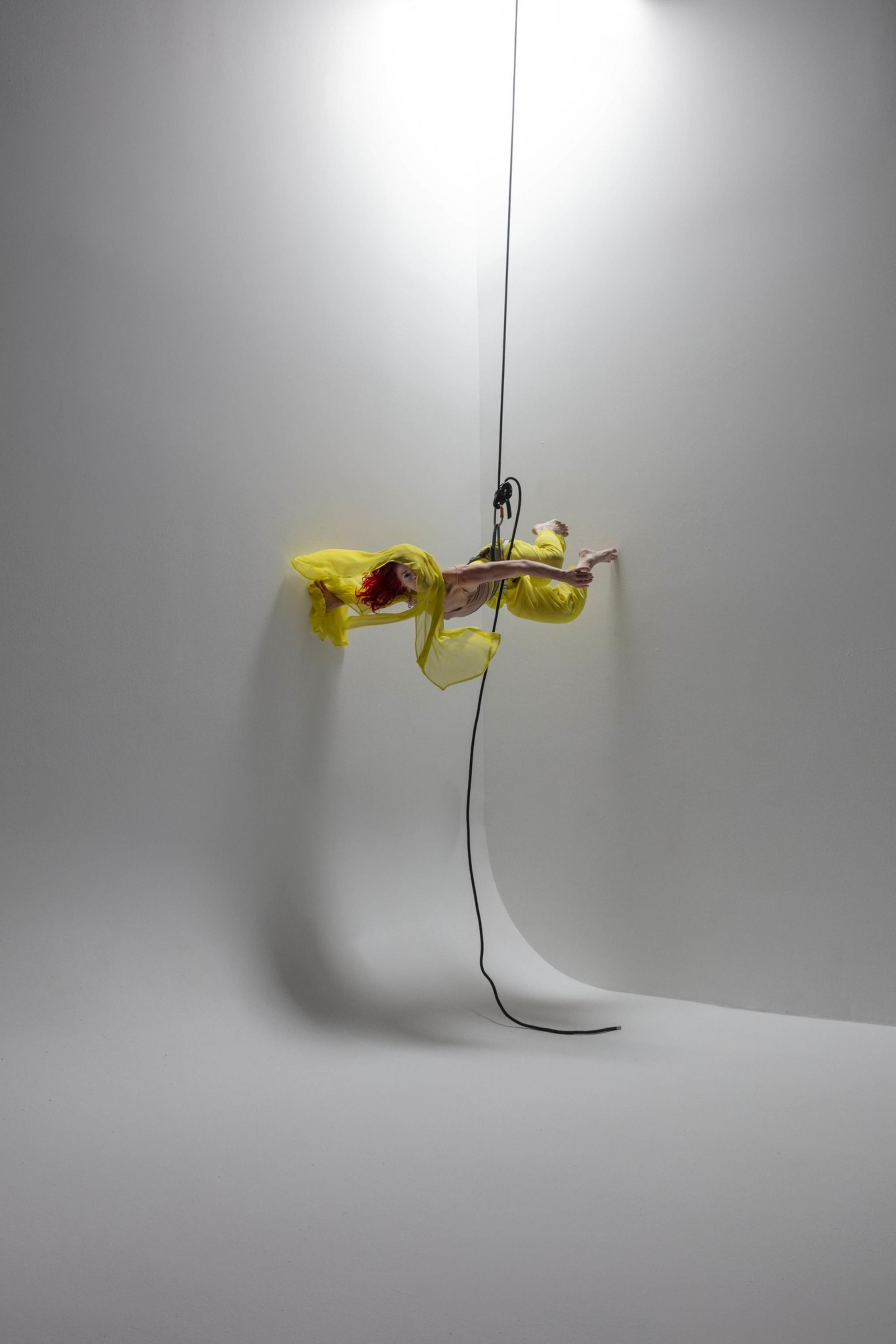 A person in a flowing yellow outfit is suspended horizontally against a white wall by ropes and harnesses, with shadows cast below.