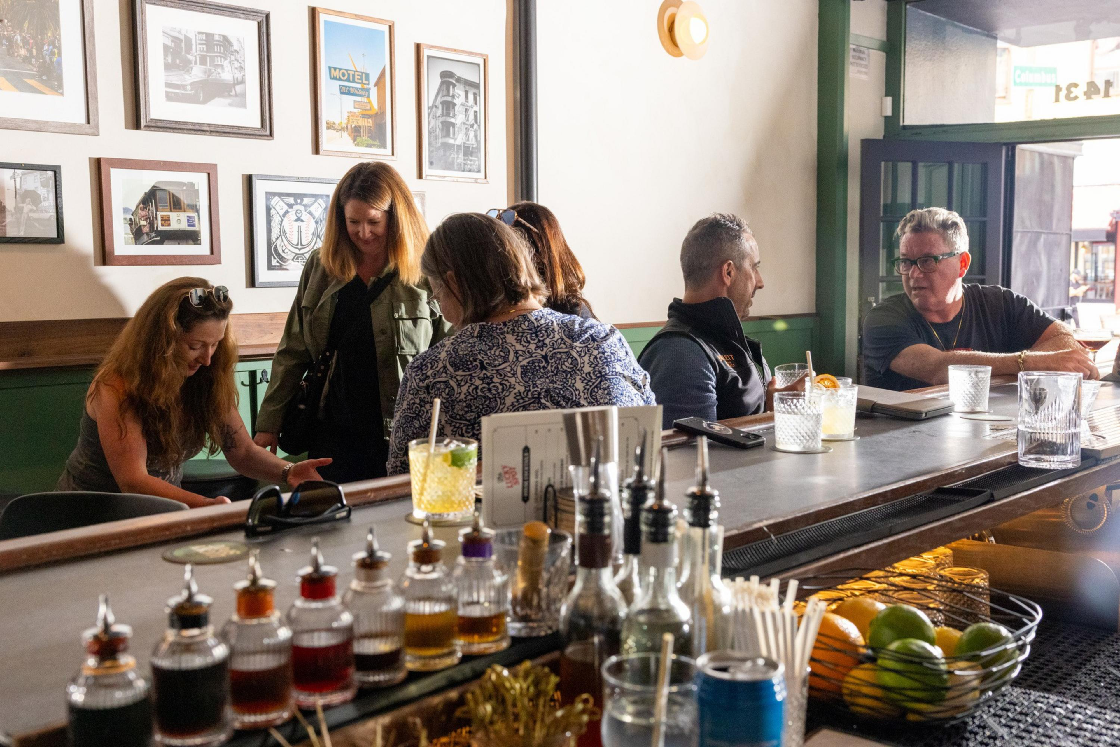 Five people sit and stand around a bar counter with drinks, bottles, and fruit, engaged in conversation in a warmly lit room with framed pictures on the wall.