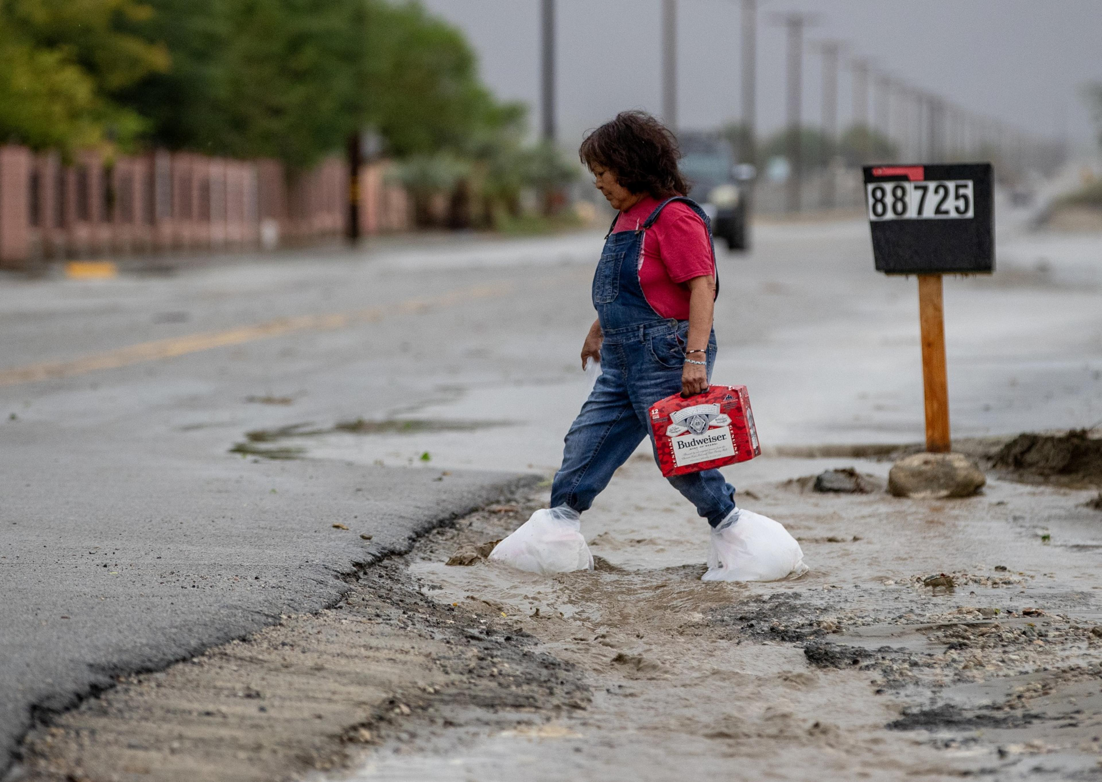 A person wearing plastic bags over their shoes walks across a wet, muddy street carrying a red Budweiser beer case.