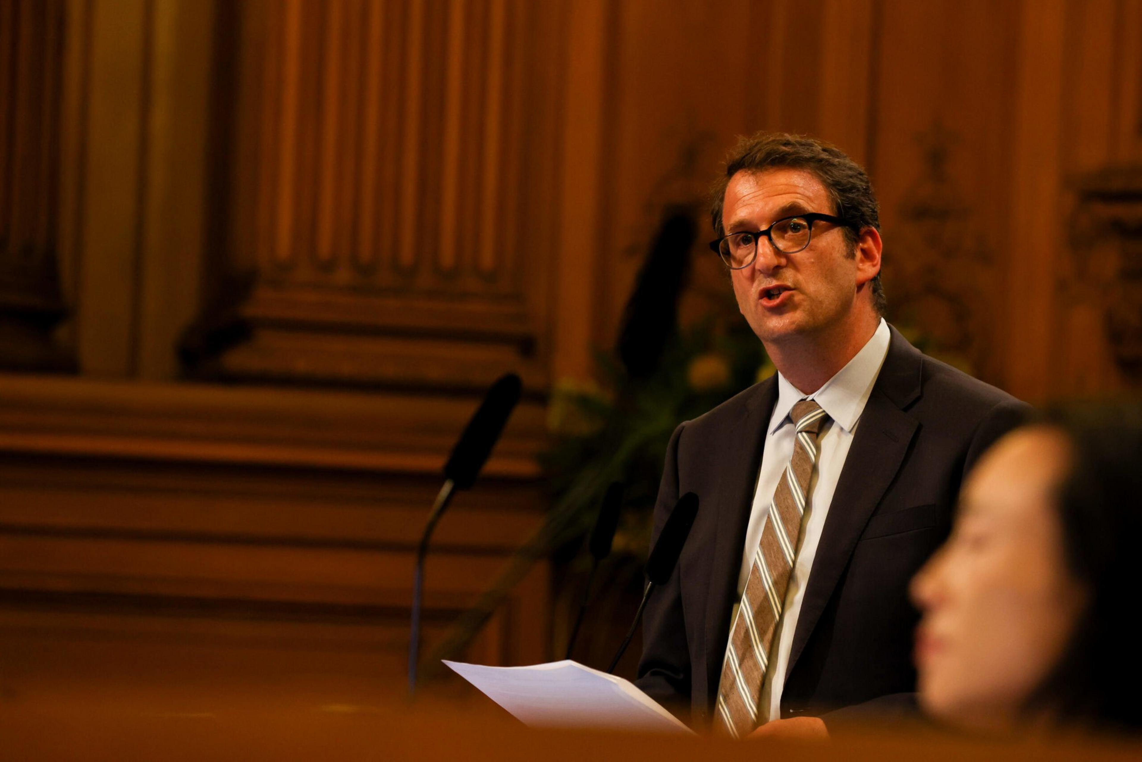 A man in a suit and striped tie is speaking while holding papers, with a blurred person in the foreground and wood-paneled walls in the background.