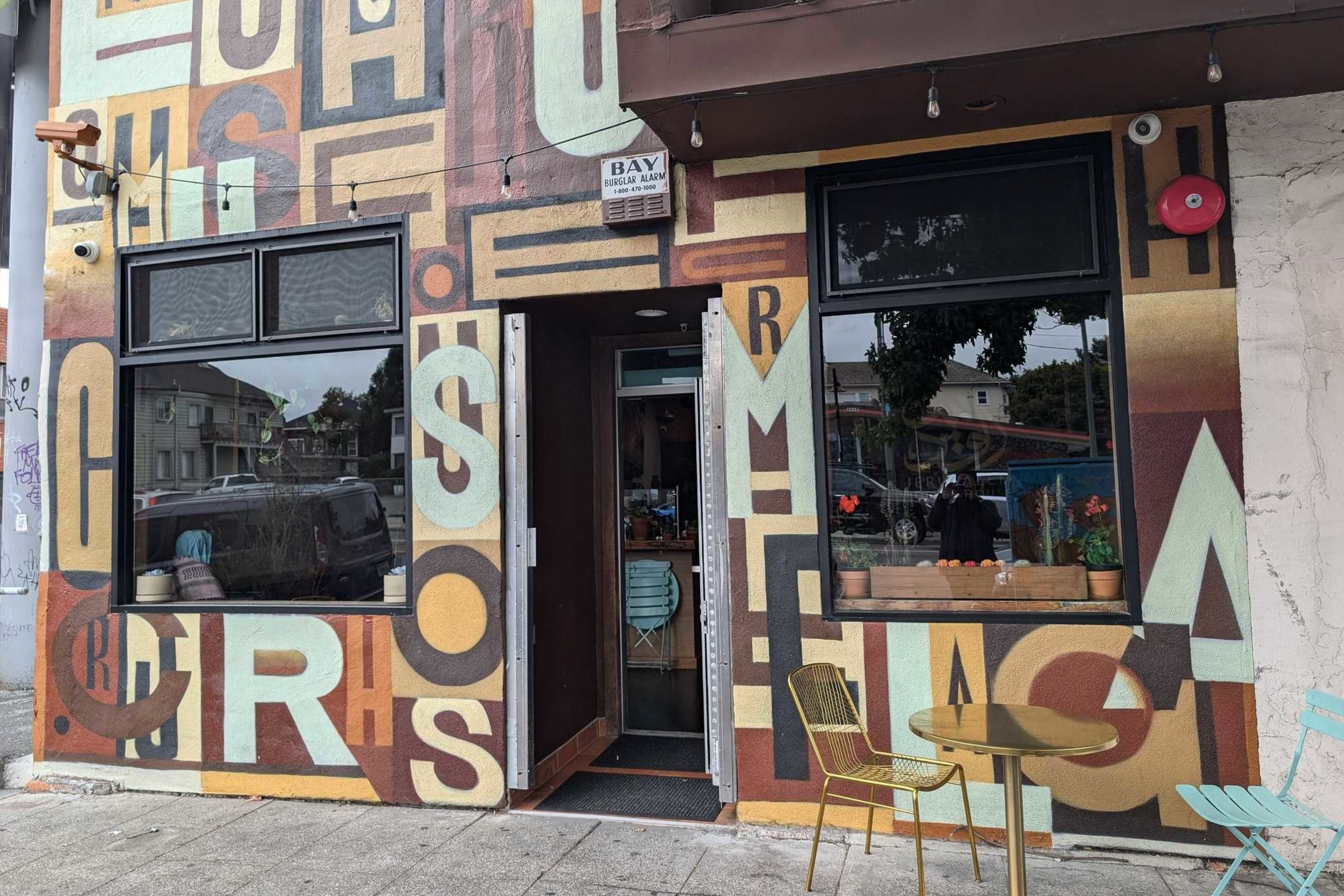 The image shows a colorful cafe exterior with large, overlapping letters painted on the walls. There are two windows and an open door, plus a small outdoor table with chairs.