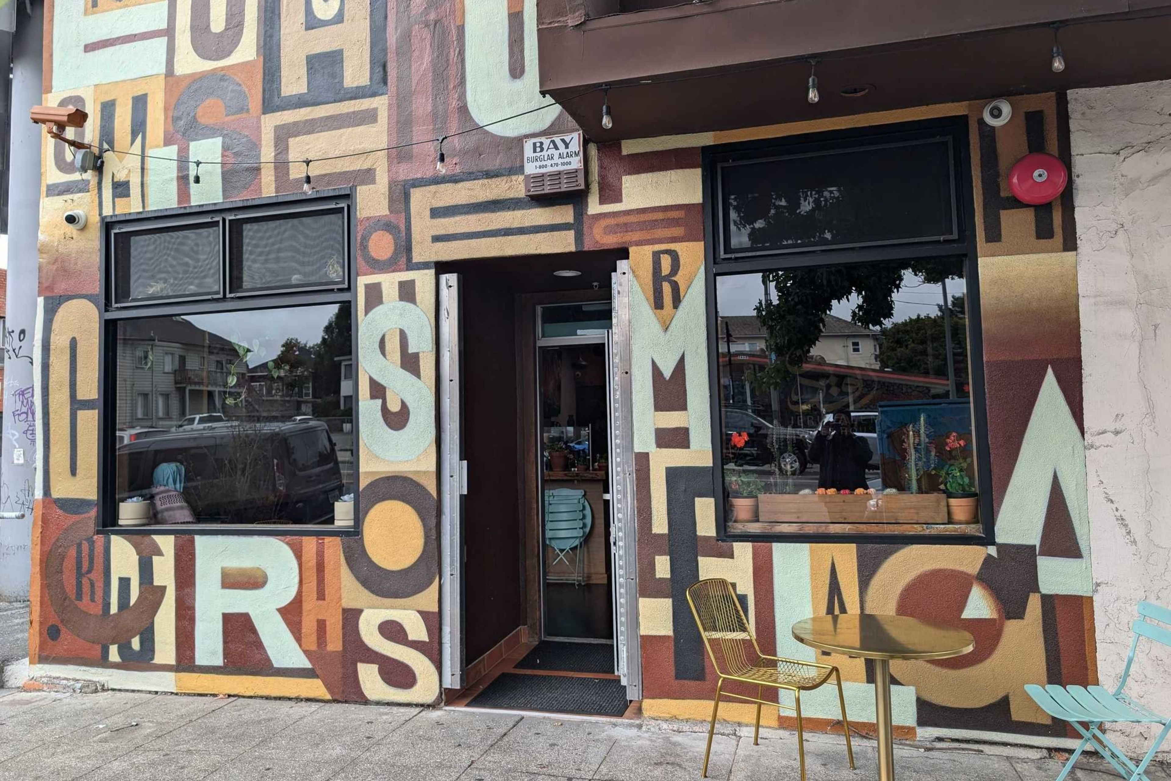 The image shows a colorful cafe exterior with large, overlapping letters painted on the walls. There are two windows and an open door, plus a small outdoor table with chairs.