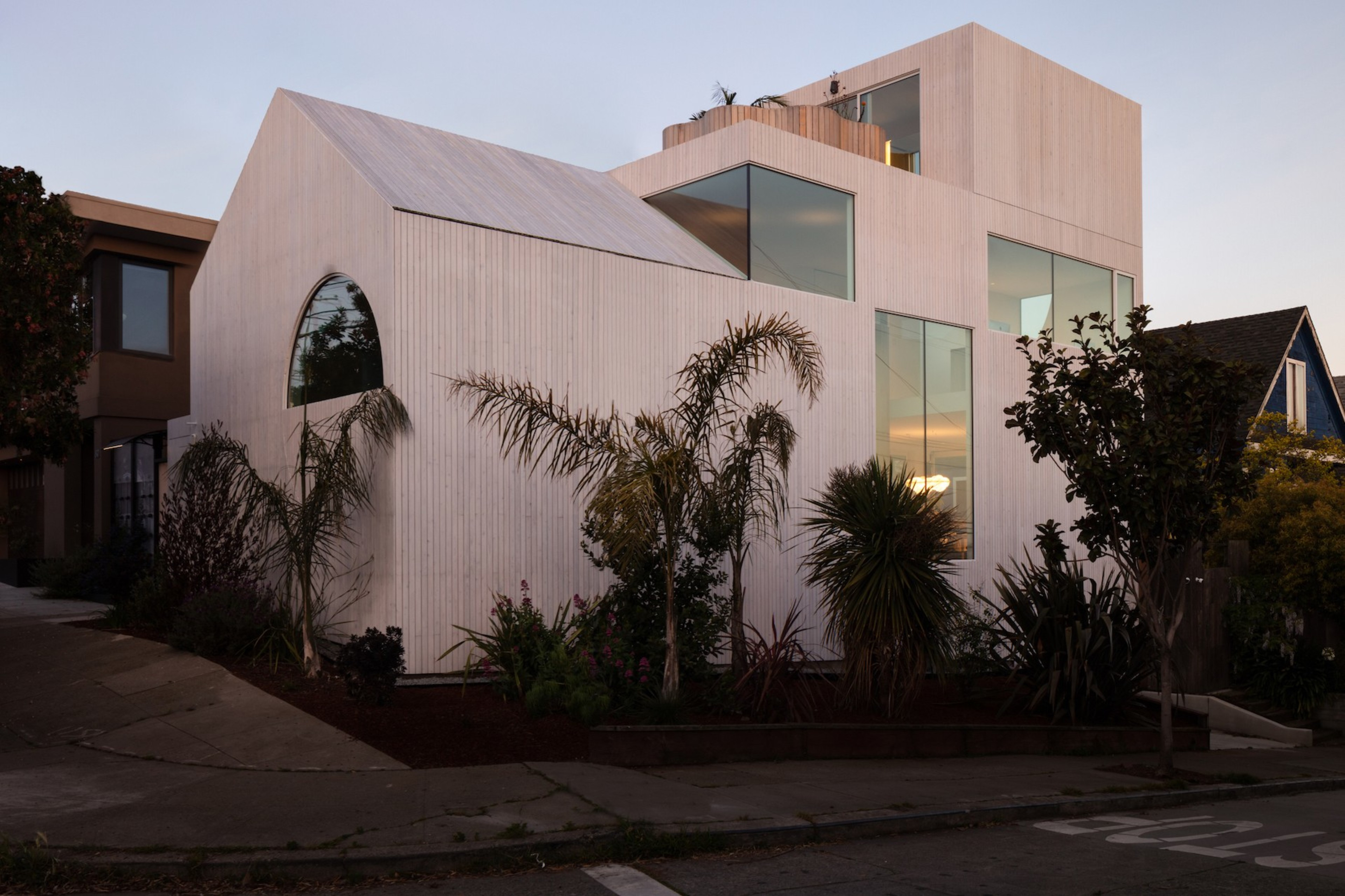 Modern house with a unique geometric design and large windows, surrounded by lush, tropical plants at twilight.