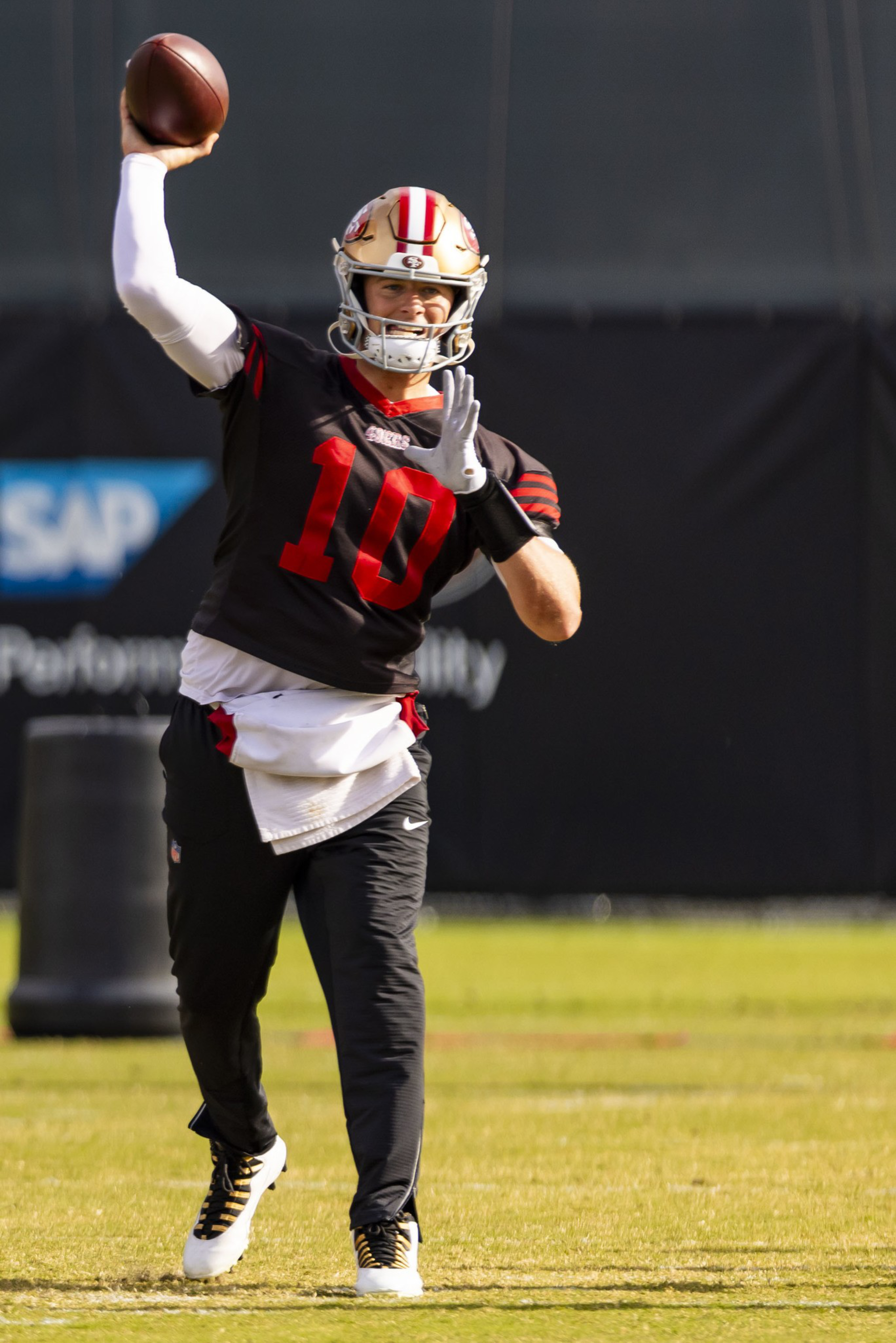 A football player wearing a black and red jersey with number 10 prepares to throw a football on a grassy field during practice.