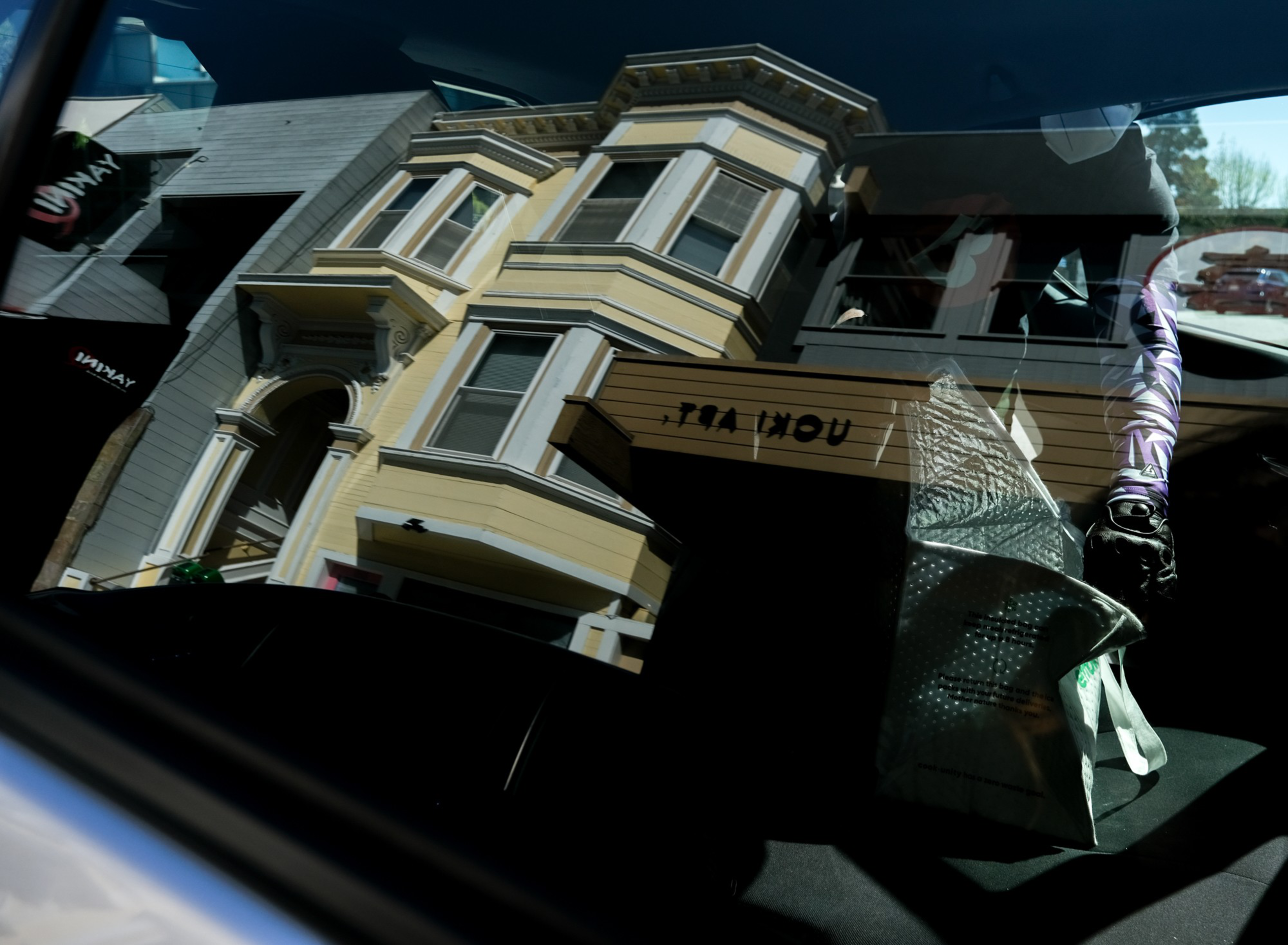A yellow Victorian-style building with ornate windows is reflected on a car window, which also shows a shiny bag and a partially visible figure inside the car.
