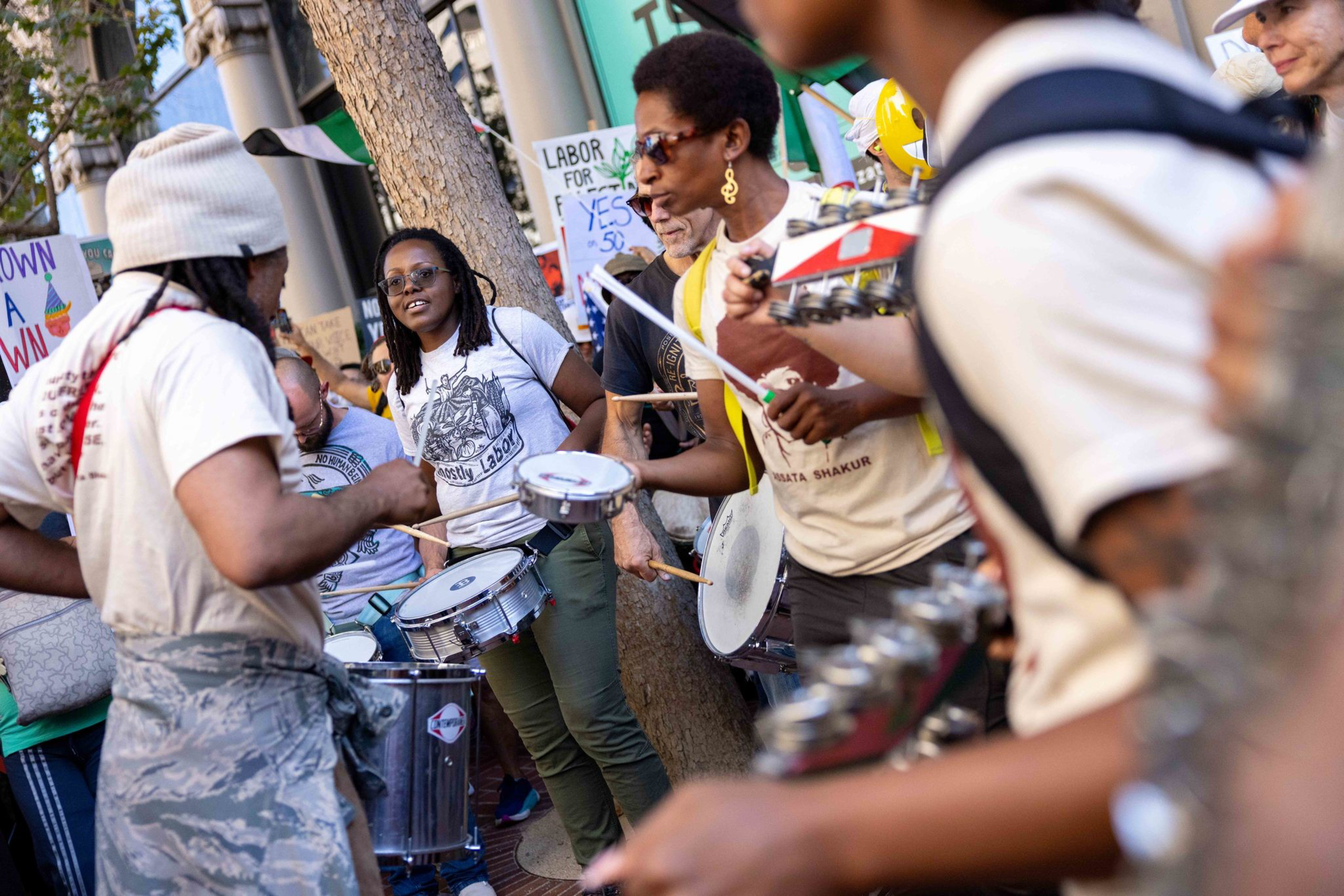 A group of people are playing drums and percussion instruments during an outdoor event with signs advocating labor rights in the background.
