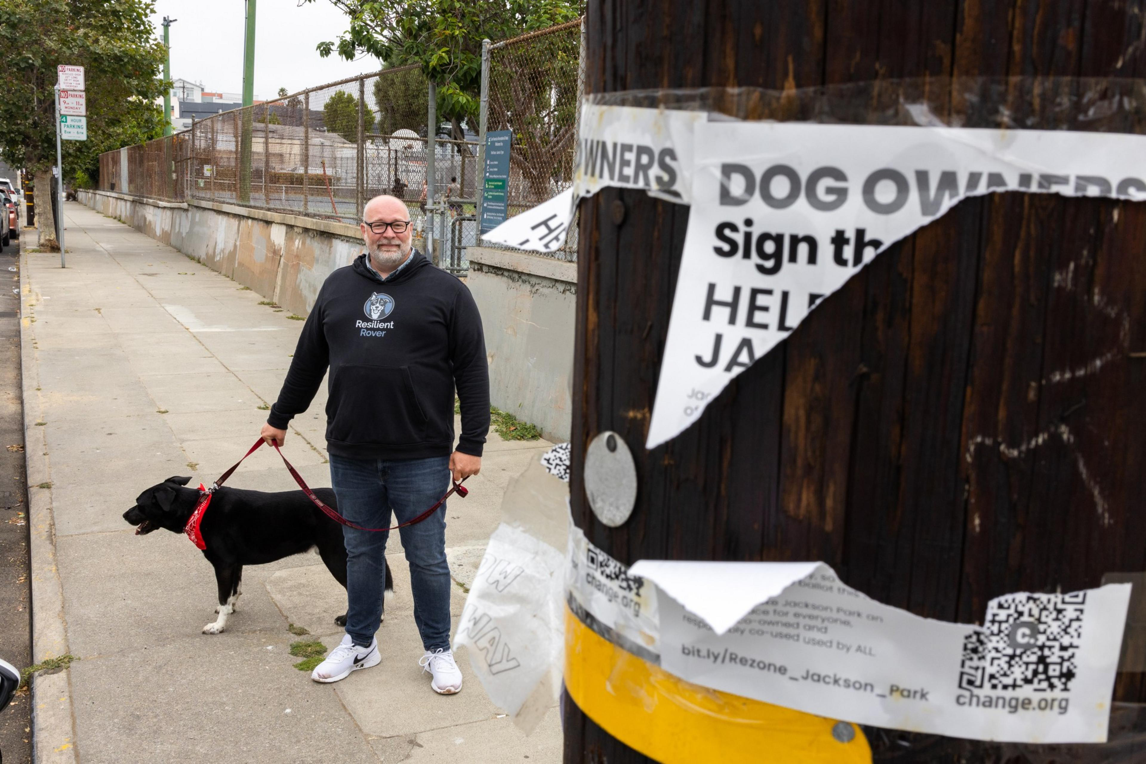 A man in a black &quot;Resilient Rover&quot; hoodie walks a black dog bandana in an urban setting near a utility pole with torn dog owner posters calling for signatures.