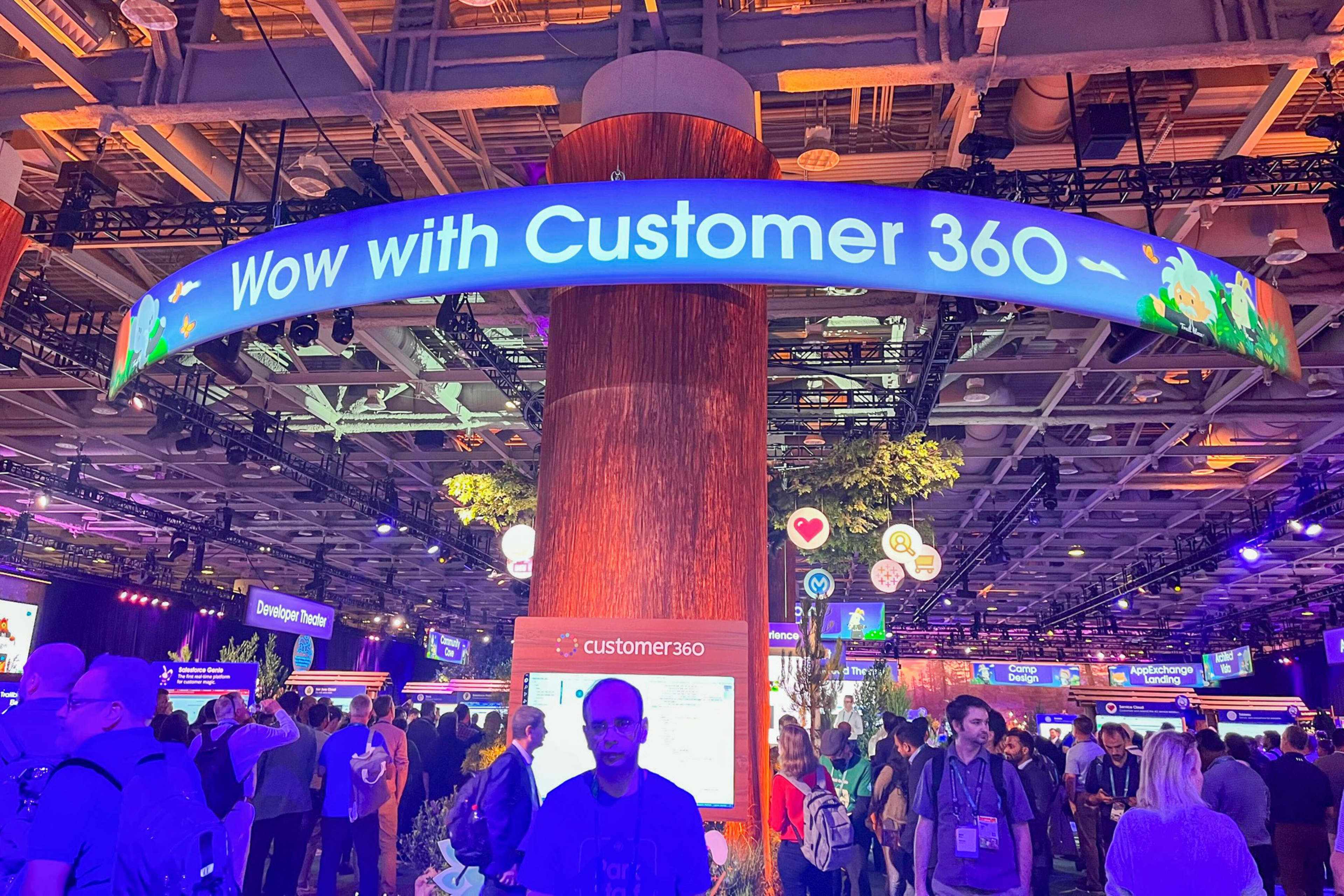 A crowded tech event features a large circular display reading "Wow with Customer 360" around a tree-like column with multiple attendees interacting and exploring booths.