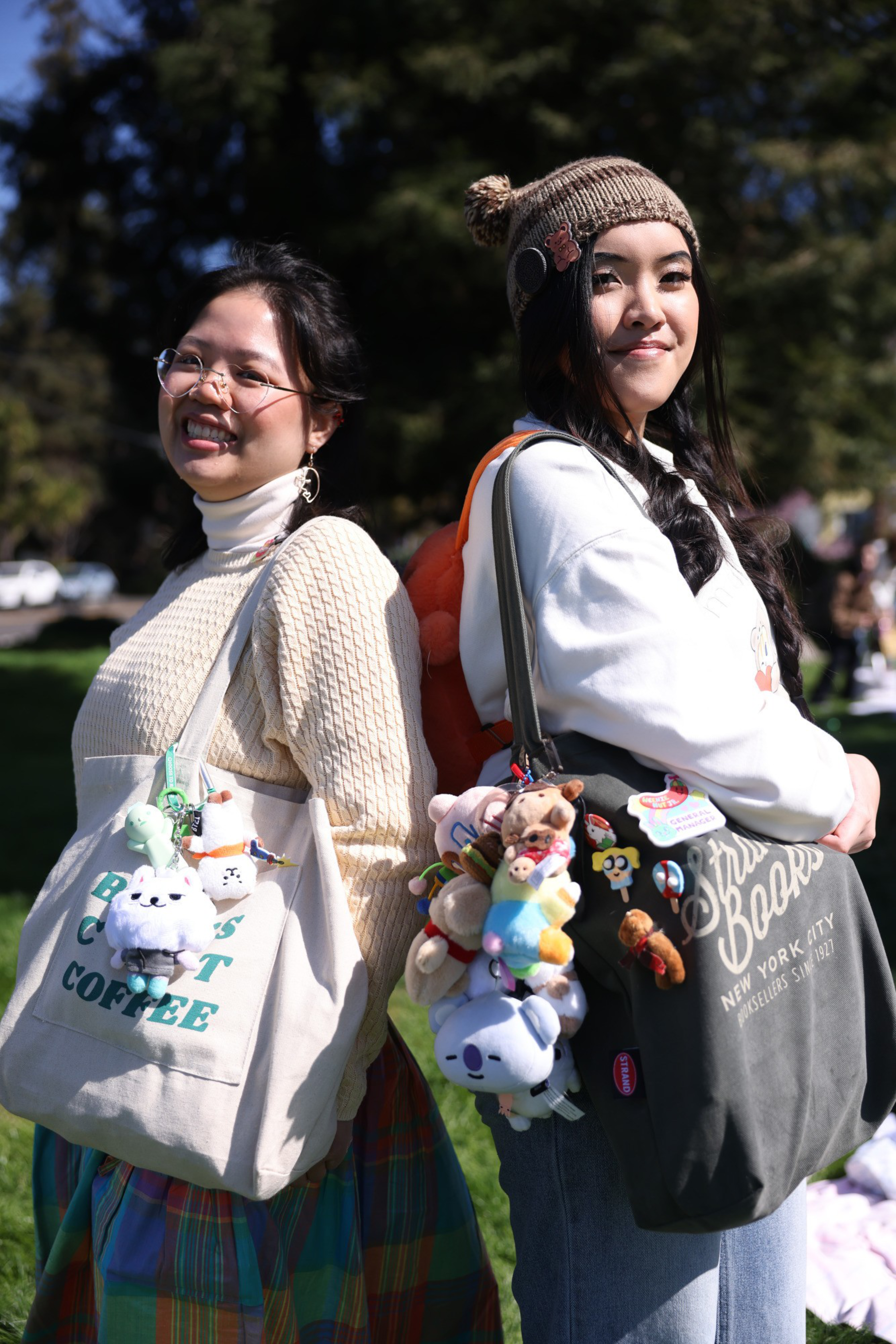 Two women stand back-to-back, smiling outdoors. They're each holding bags adorned with various cute plush charms, and one wears a knit hat with decorative pins.