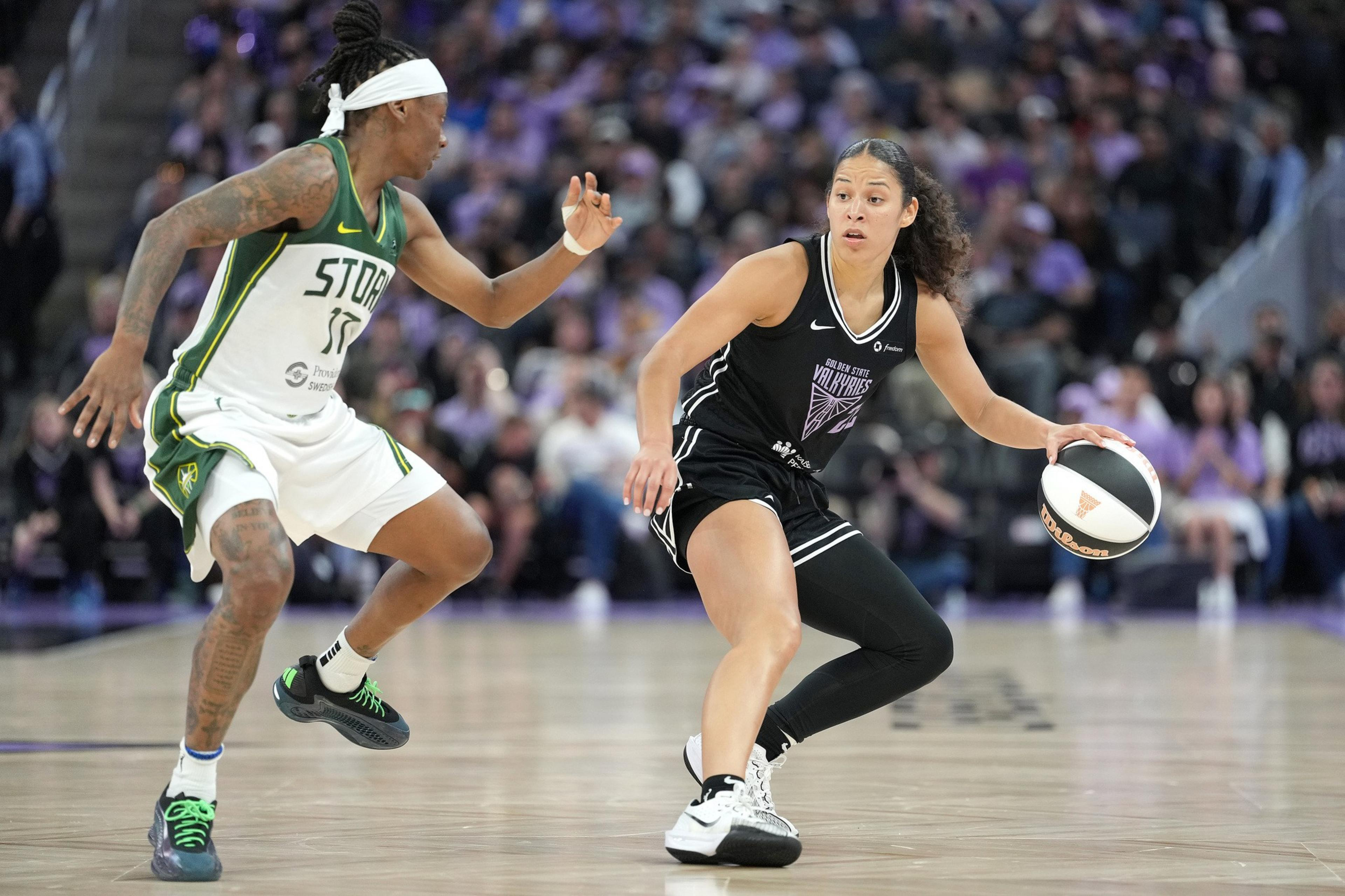 Two basketball players are on a court. One is in a green and white uniform, defending the other, who is dribbling the ball in a black uniform.