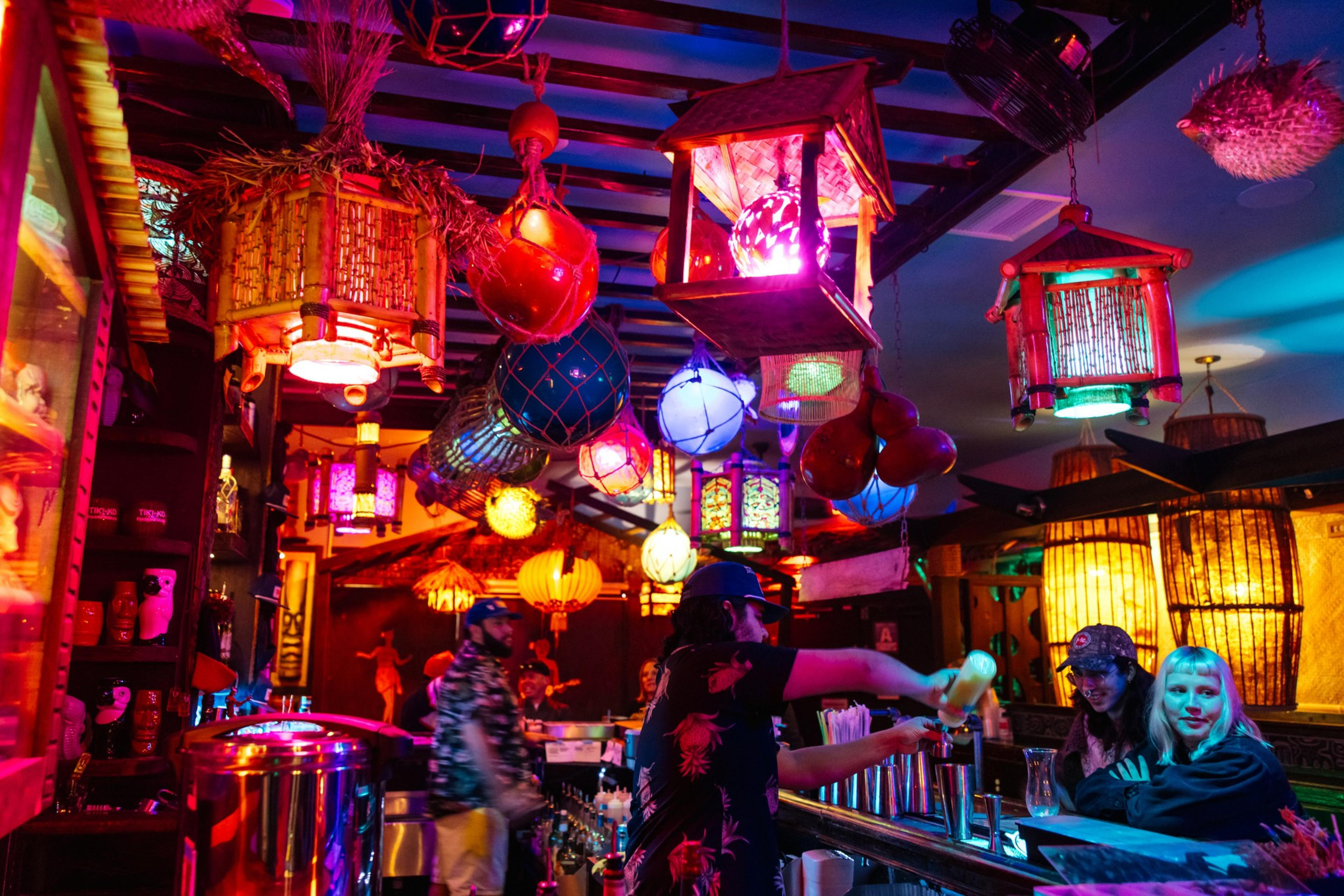 The image shows a vibrant bar with multicolored lanterns and lights hanging from the ceiling. People are seated at the bar, with bottles and glasses visible.