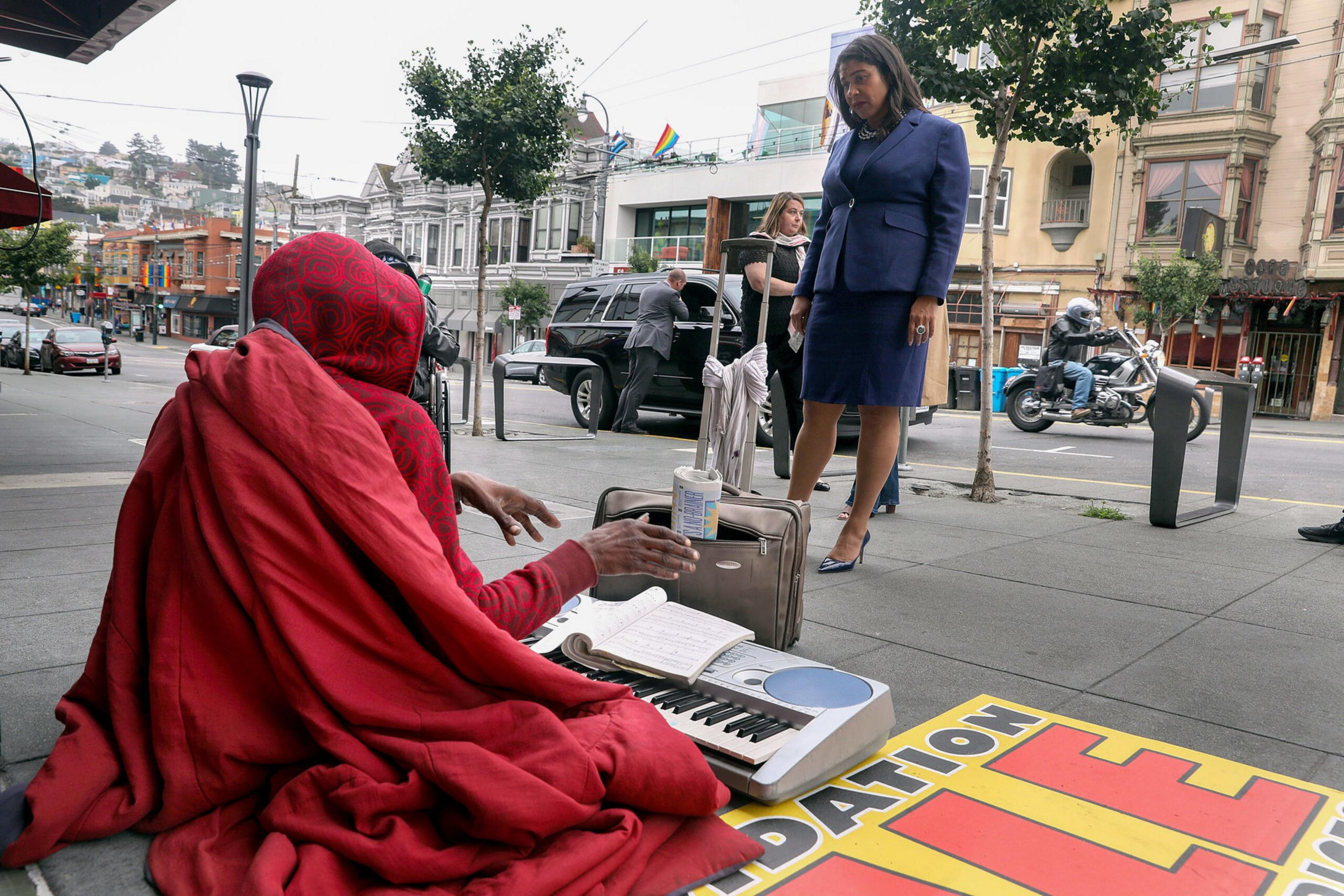 Mayor Lond Breed stands beside a homeless person sitting on the sidewalk
