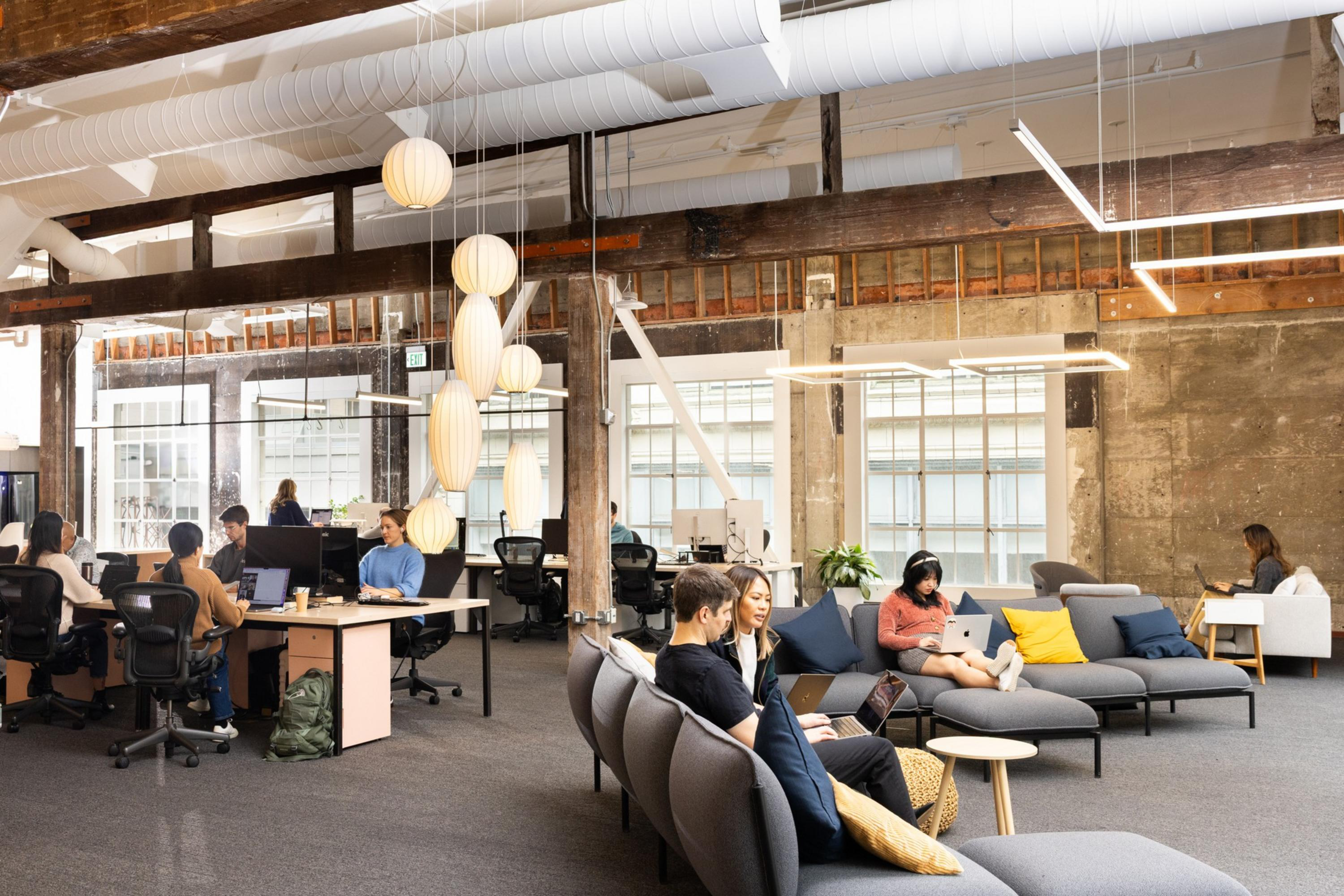 People work on laptops and computers in a bright, open office with modern furniture, large windows, and hanging pendant lights.