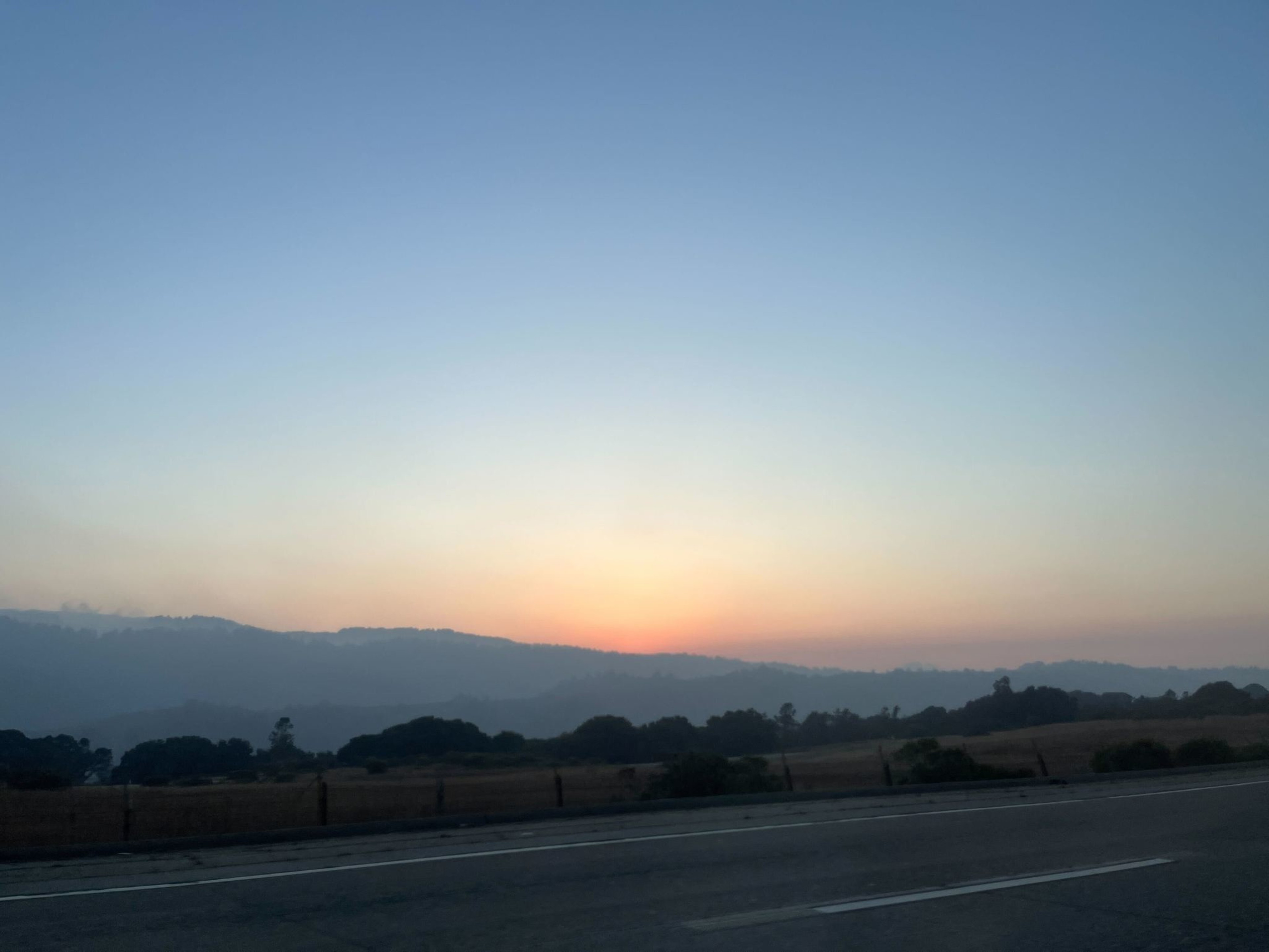 A peaceful sunset with a clear sky above layered hills. The horizon glows softly orange, and a highway runs along the bottom of the image.