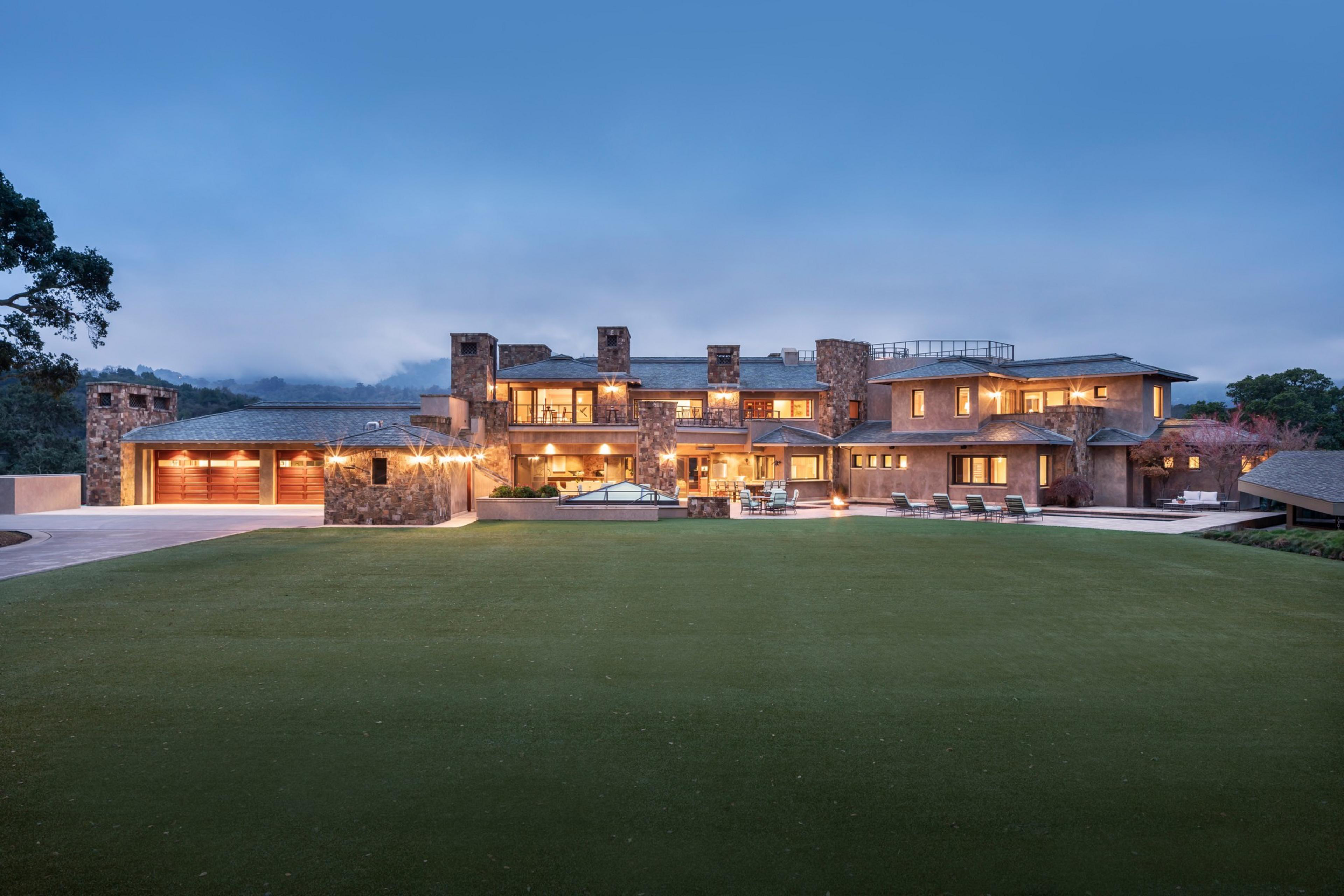 A large, luxurious house with stone exterior walls, lit warmly, stands in front of a vast, manicured lawn with lawn chairs and outdoor lighting at dusk.