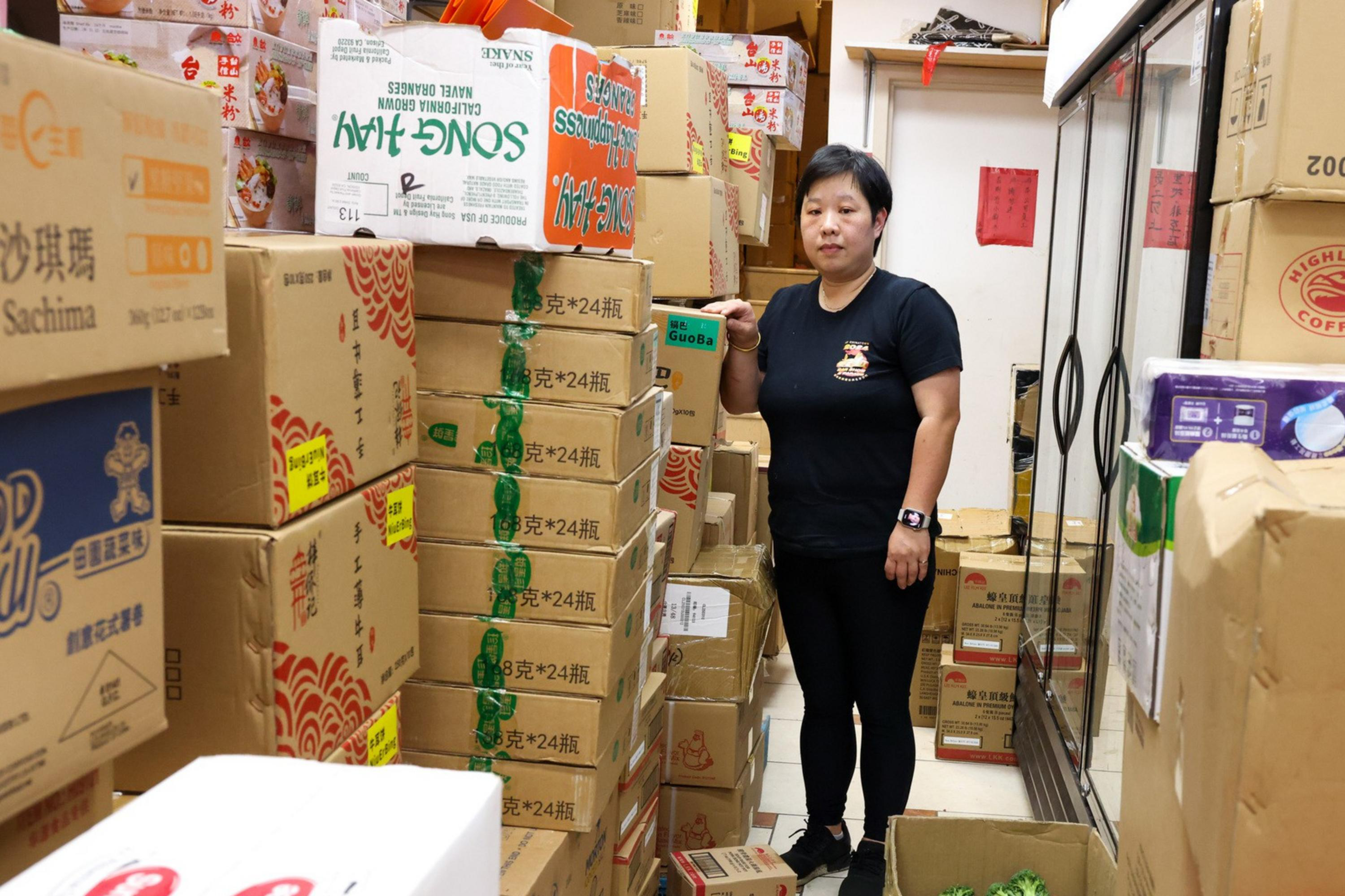 A woman stands in a narrow storage area filled with stacked cardboard boxes, many labeled with Asian brands. She wears black attire and a smartwatch.