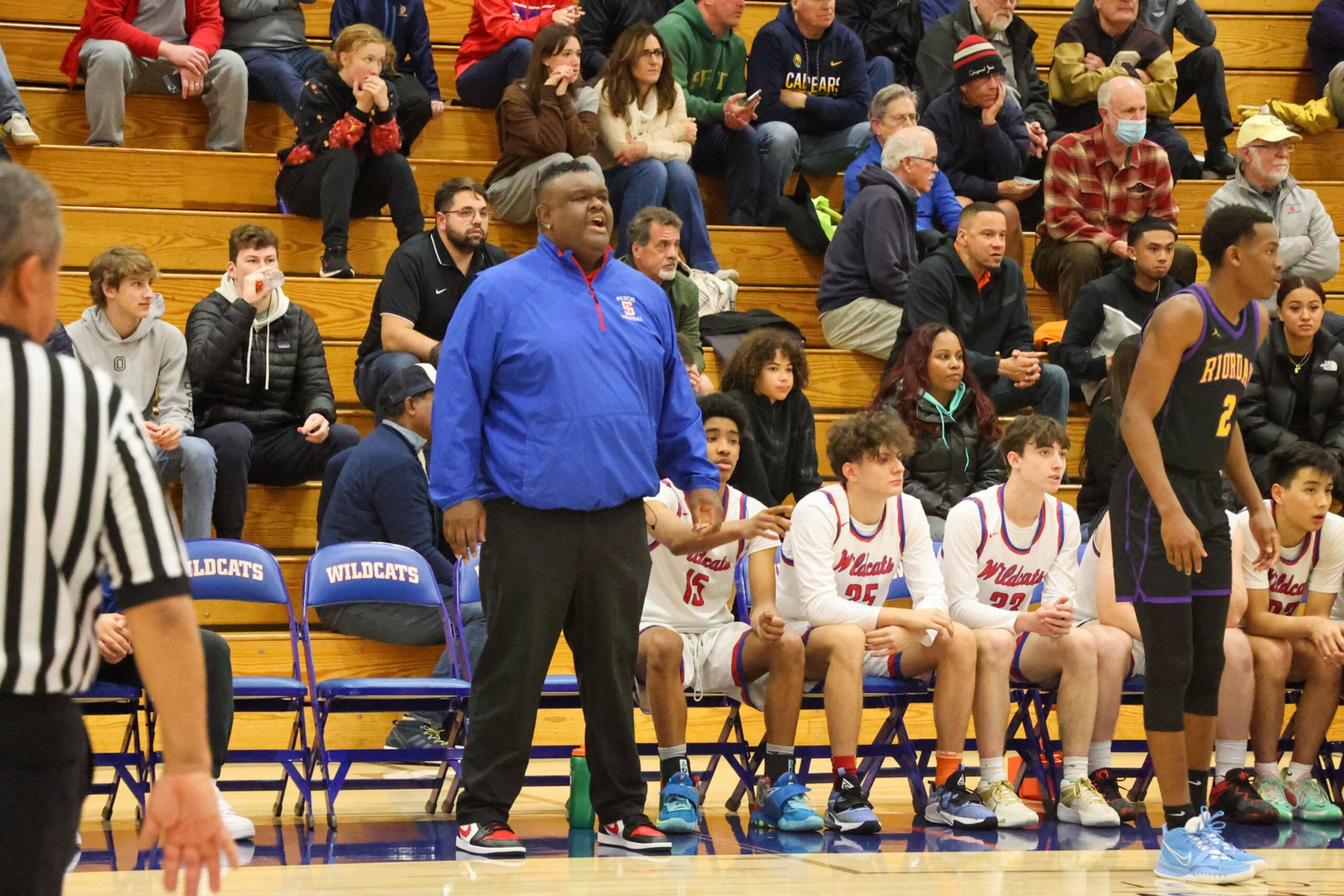 A basketball coach in a blue jacket stands shouting near seated players in white uniforms labeled Wildcats, with spectators watching from wooden bleachers.