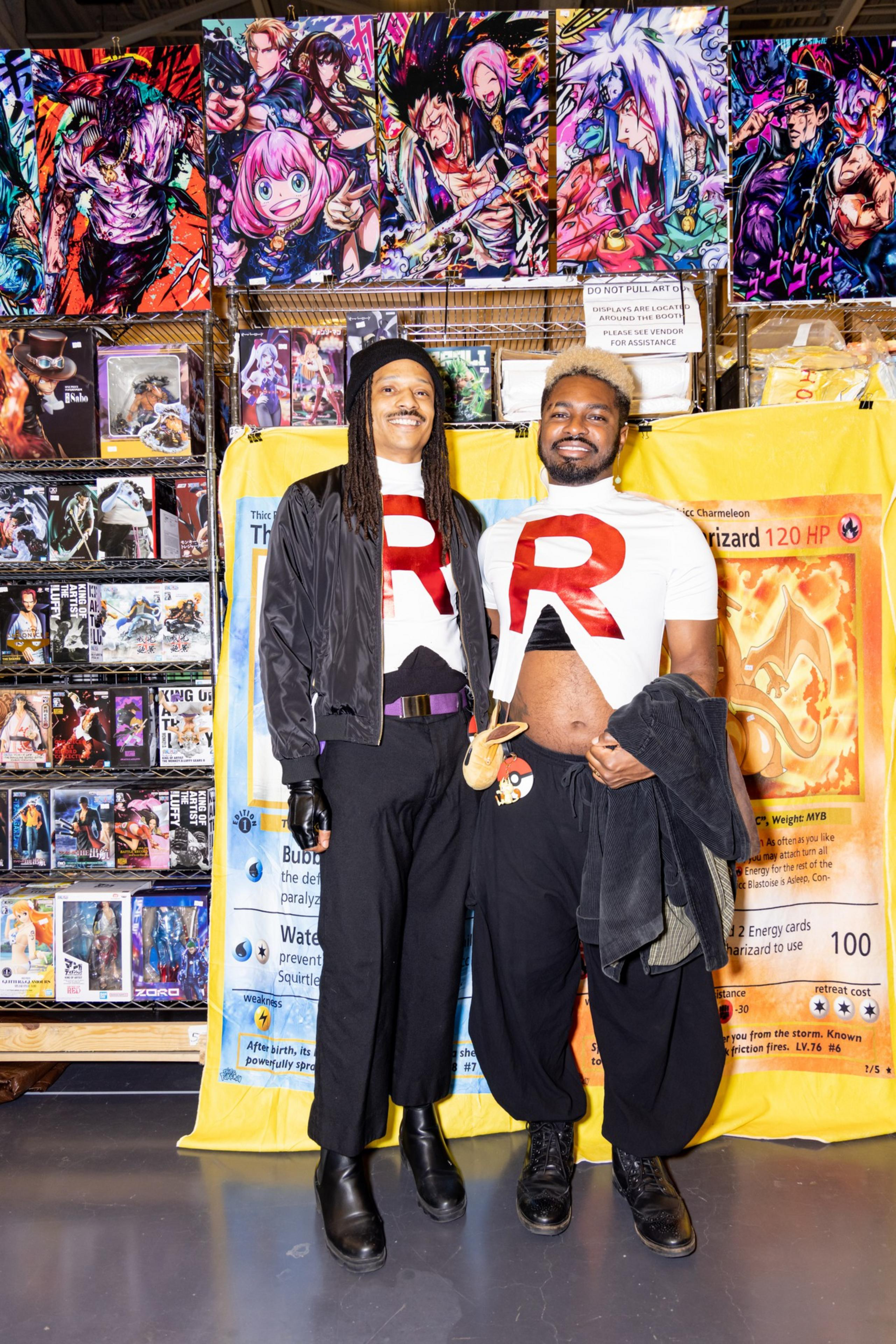 Two people in matching &quot;R&quot; shirts pose in front of anime art and collectible figures, with a giant Charizard card backdrop. They smile, exuding fun and excitement.