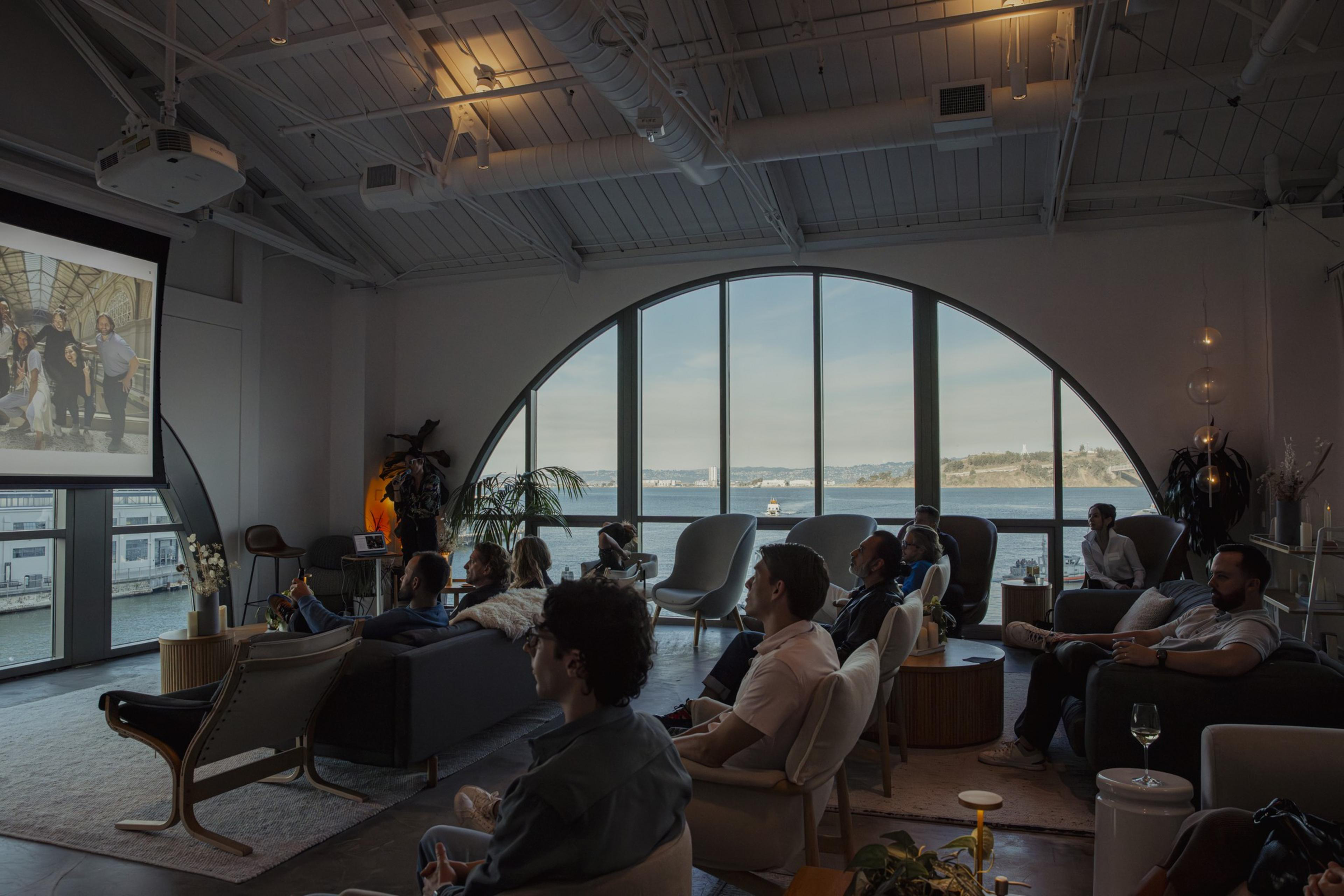 A group of people are seated in a modern room with large arched windows overlooking a body of water. They are watching a projected image on a screen.