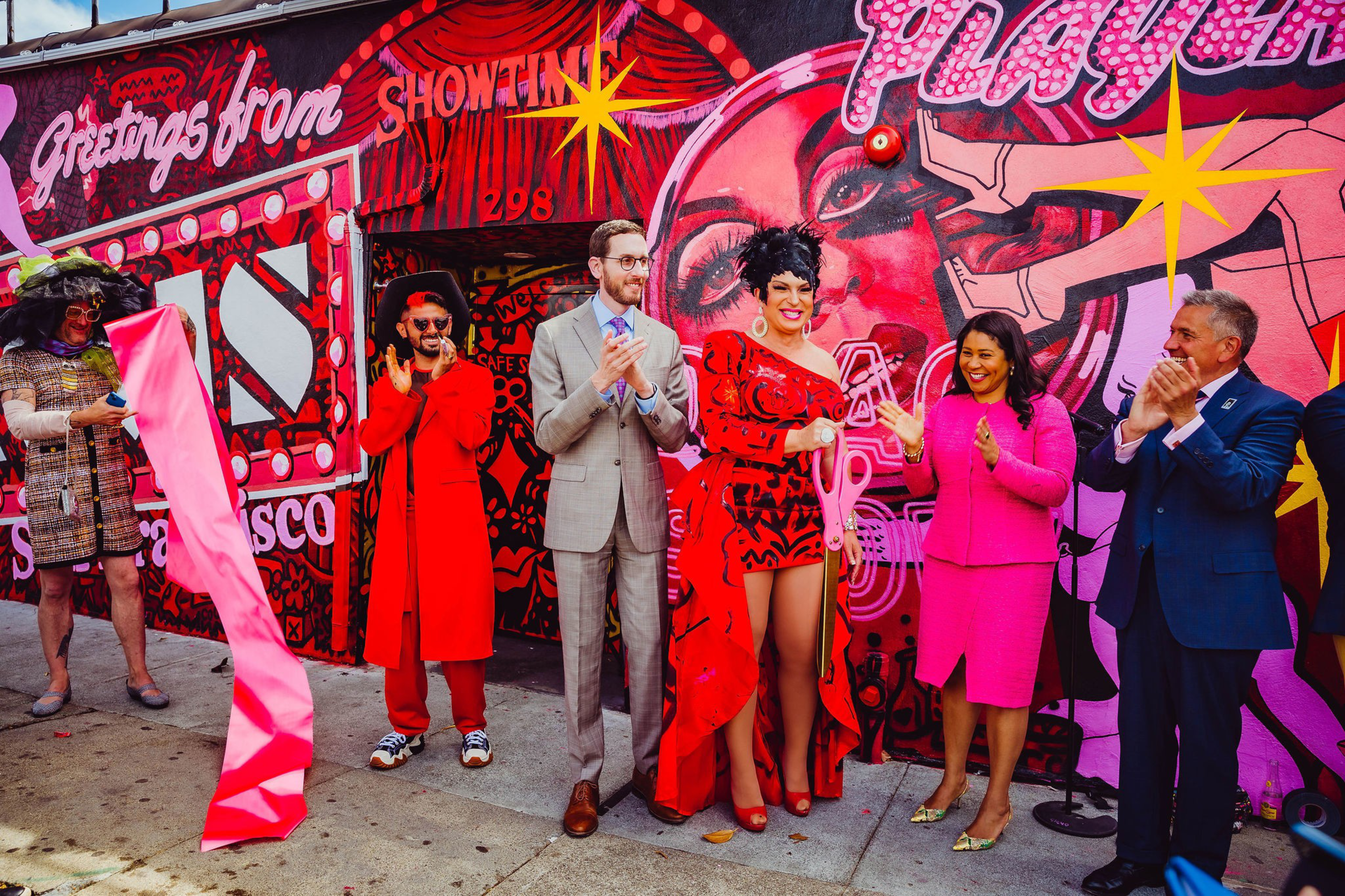 Five people stand and applaud in colorful clothing in front of a vibrant mural reading "Greetings from San Francisco" and "Showtime."