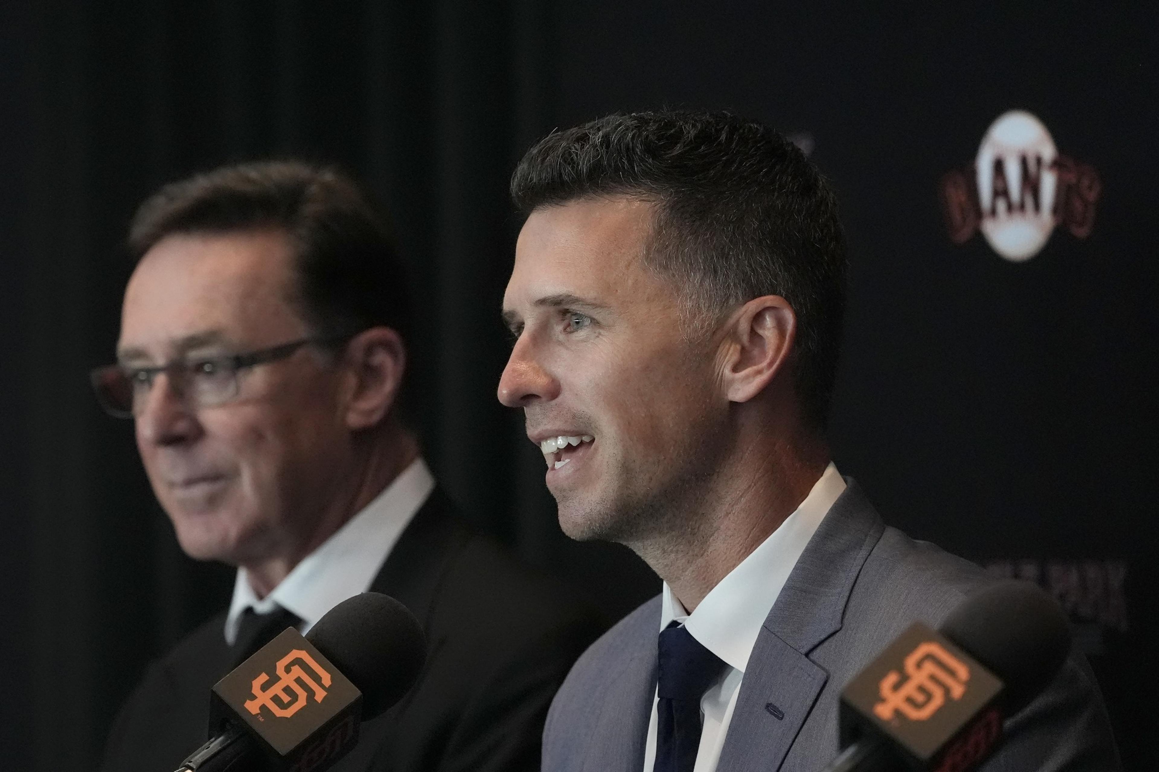 Two men in suits are speaking at a press conference. One smiles while speaking into a microphone with an orange logo, and the other looks on.