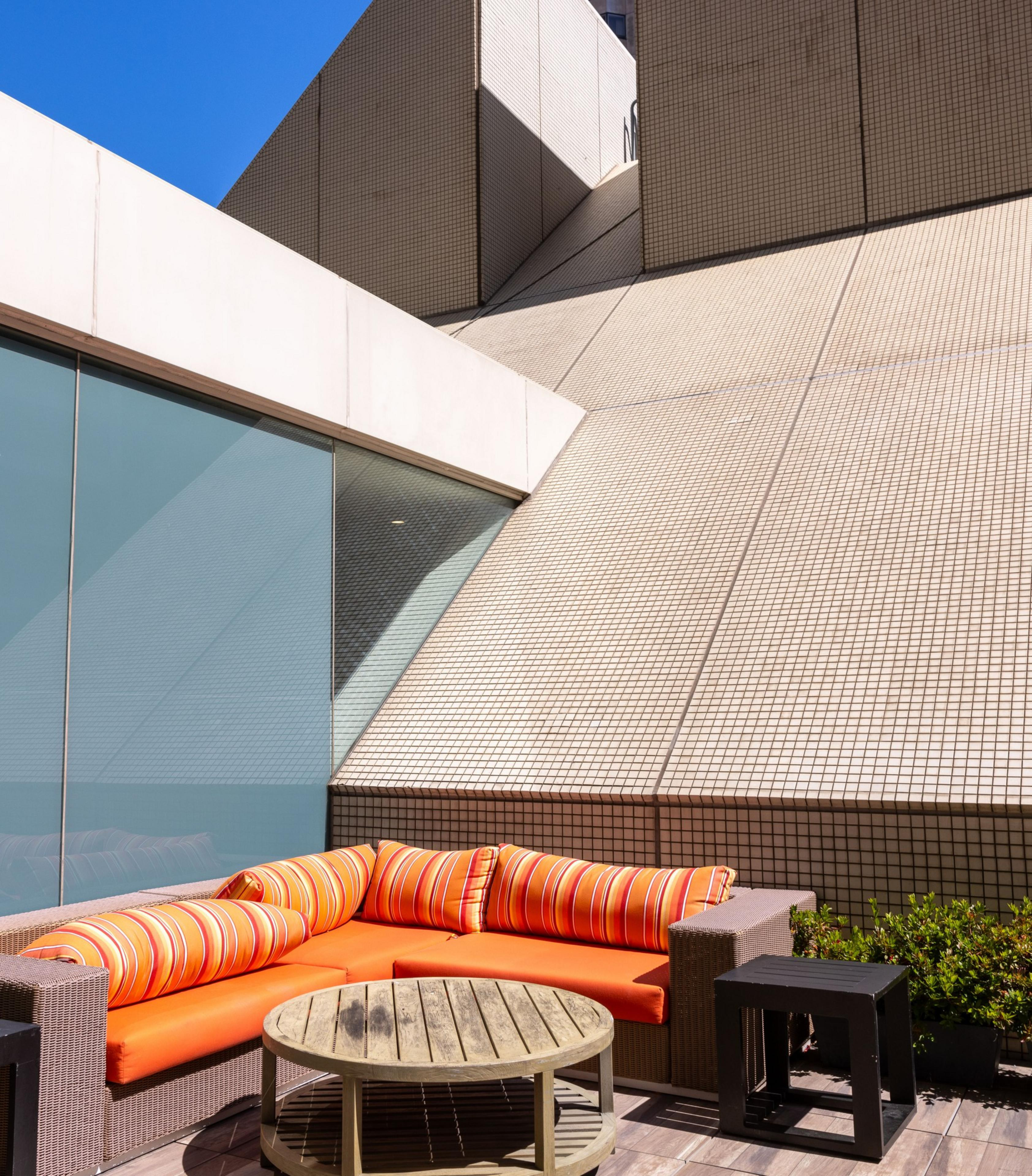 The image shows an outdoor seating area with an orange cushioned sectional sofa and striped pillows. It is set against angular, tiled building walls under a clear blue sky.