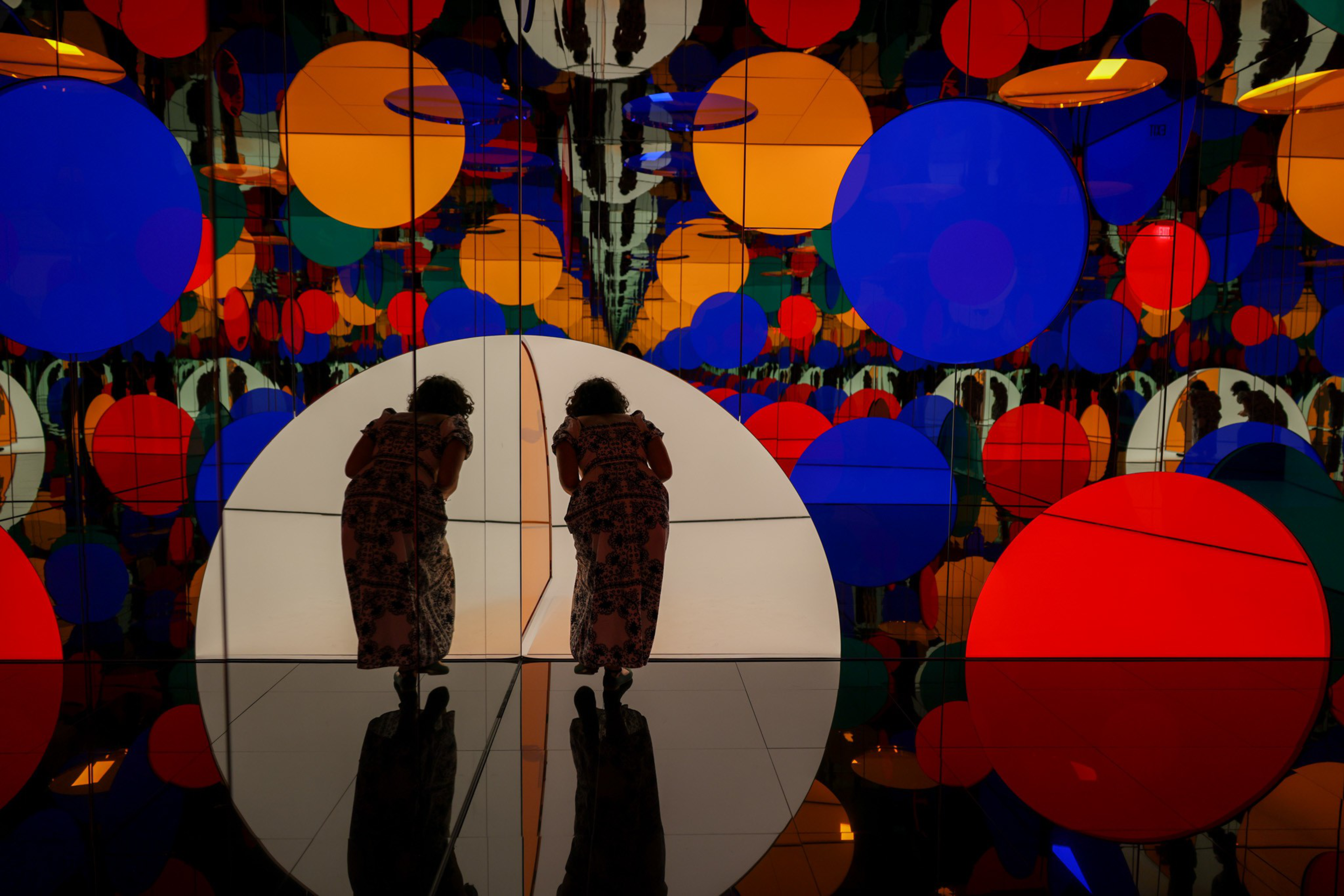 A person in a patterned dress leans forward, reflected twice amid large colorful circles on mirrored walls and floor.