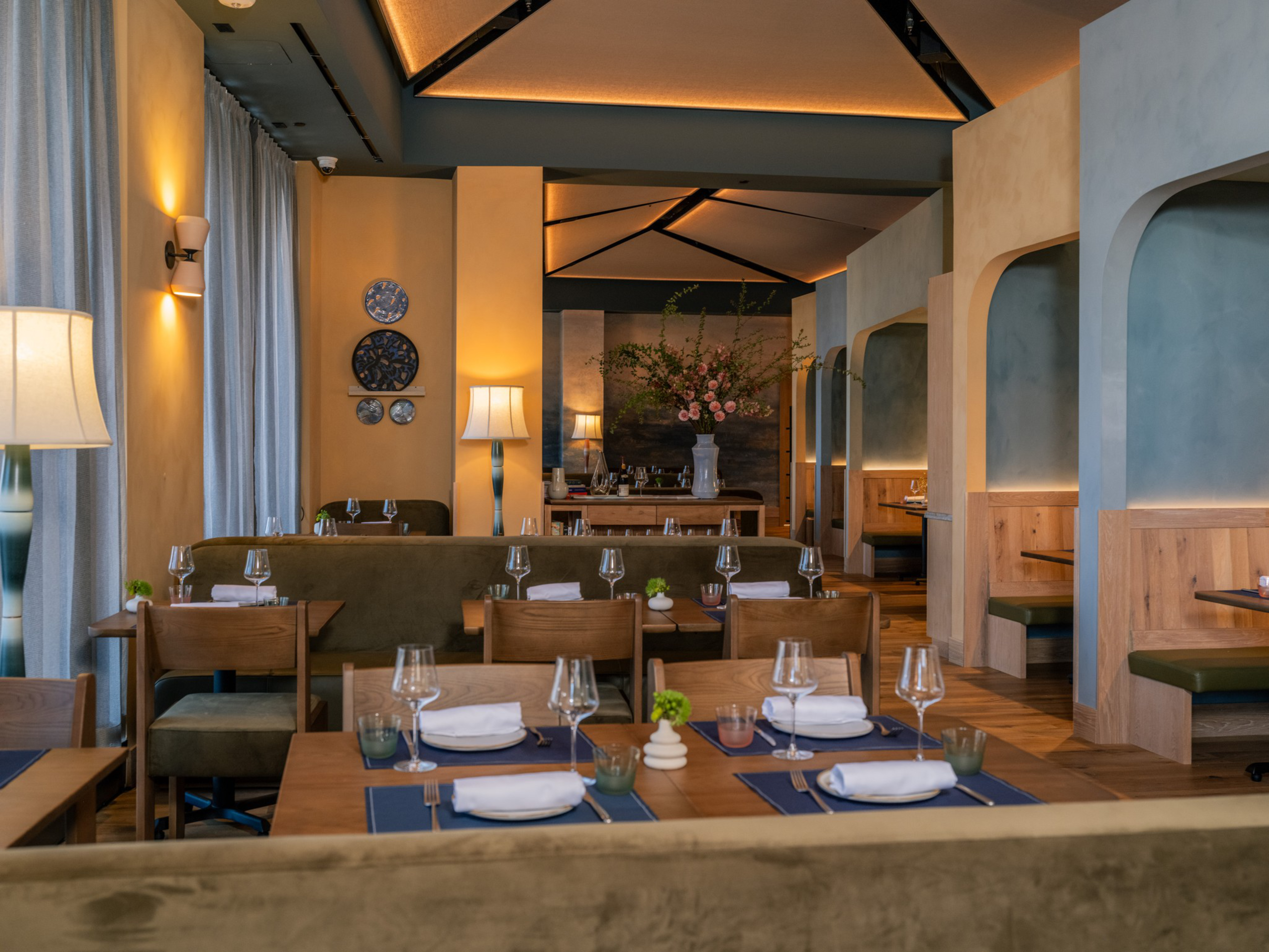 A modern restaurant with wooden tables set with white napkins and wine glasses, green cushioned seats, decorative plates on the wall, and soft lighting.