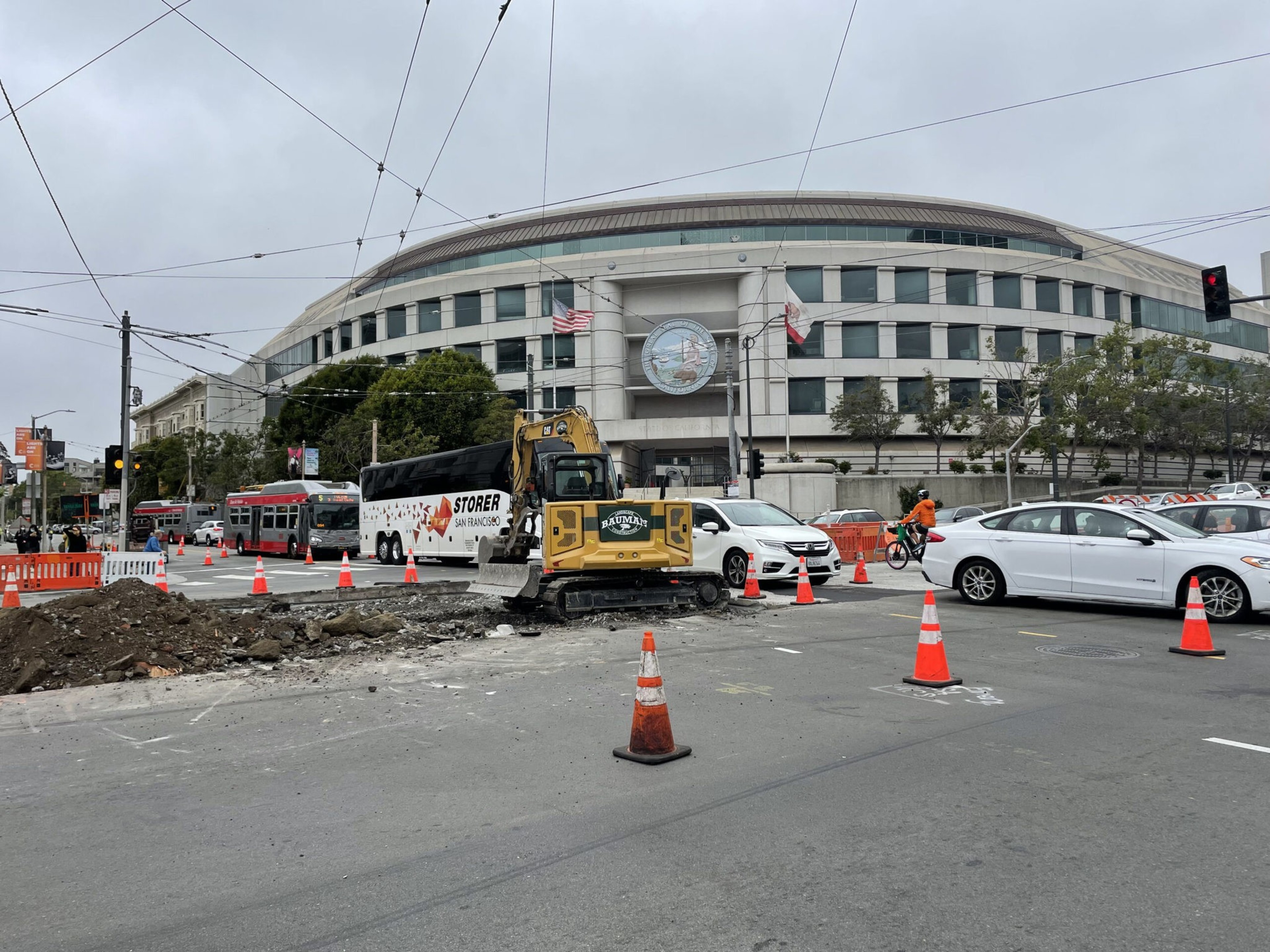 A construction site with an excavator and orange cones blocks part of a busy street, with cars, a bus, and a cyclist near a large curved building.