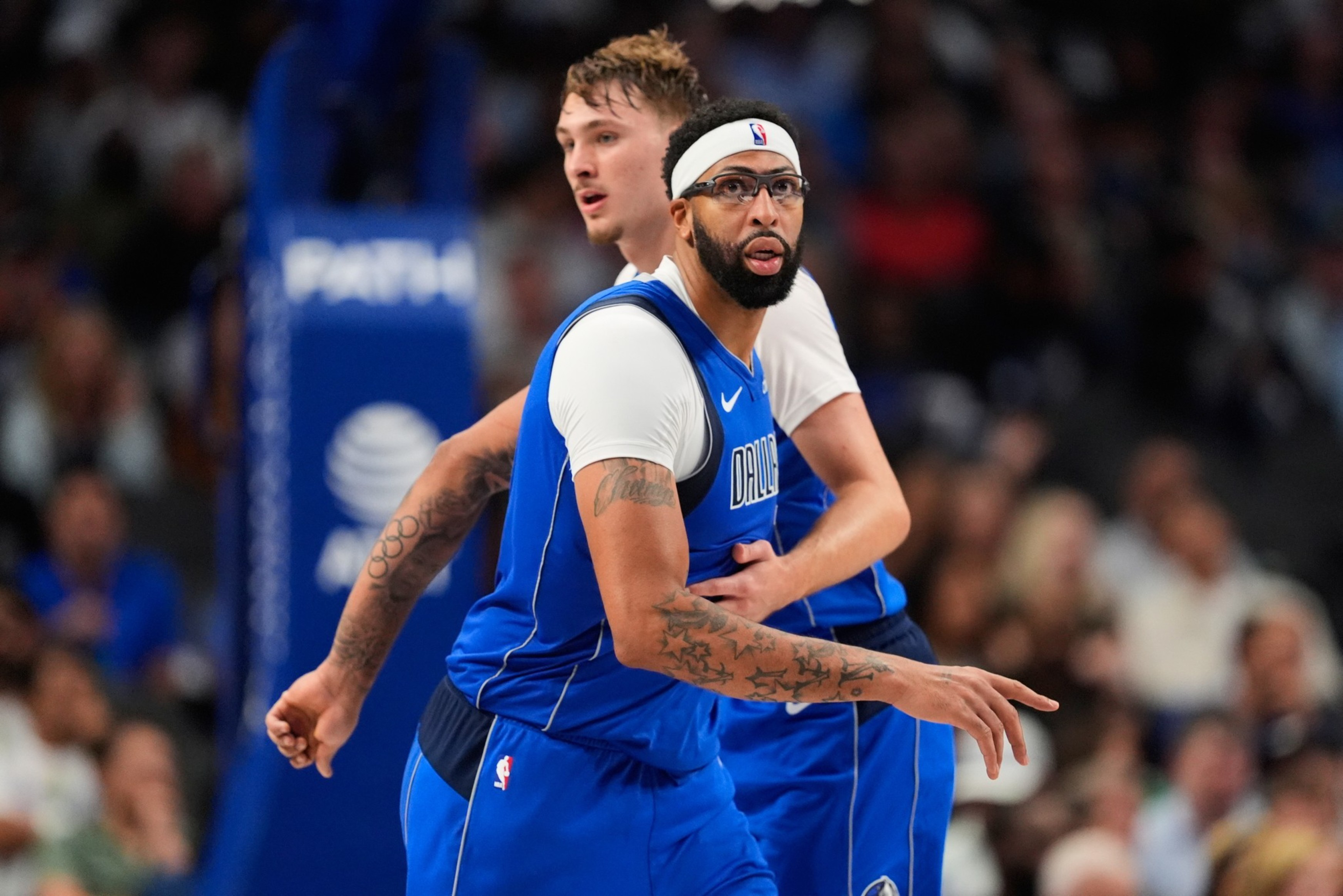 A basketball player wearing a blue Dallas Mavericks uniform and white headband is closely guarded by another player during a game.