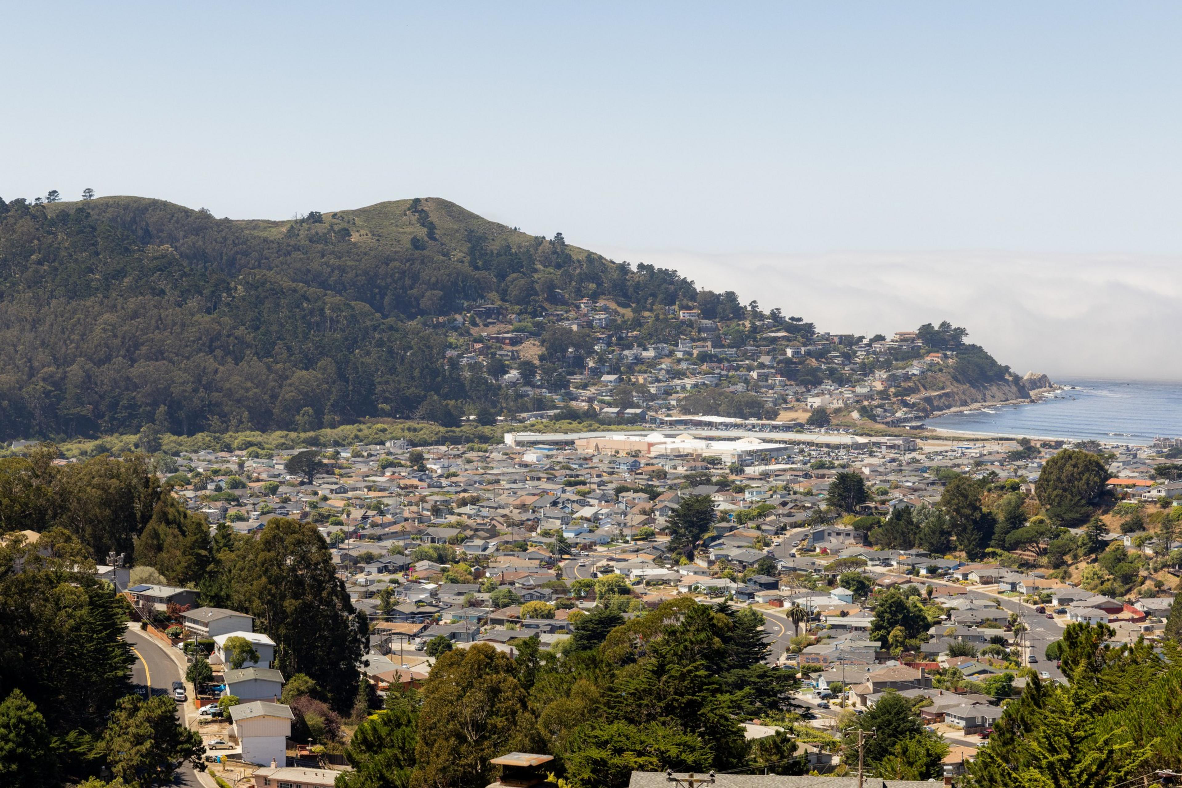 A coastal town with densely packed houses sits below rolling, tree-covered hills, and near a rocky shoreline. The sea is visible with low clouds on the horizon.