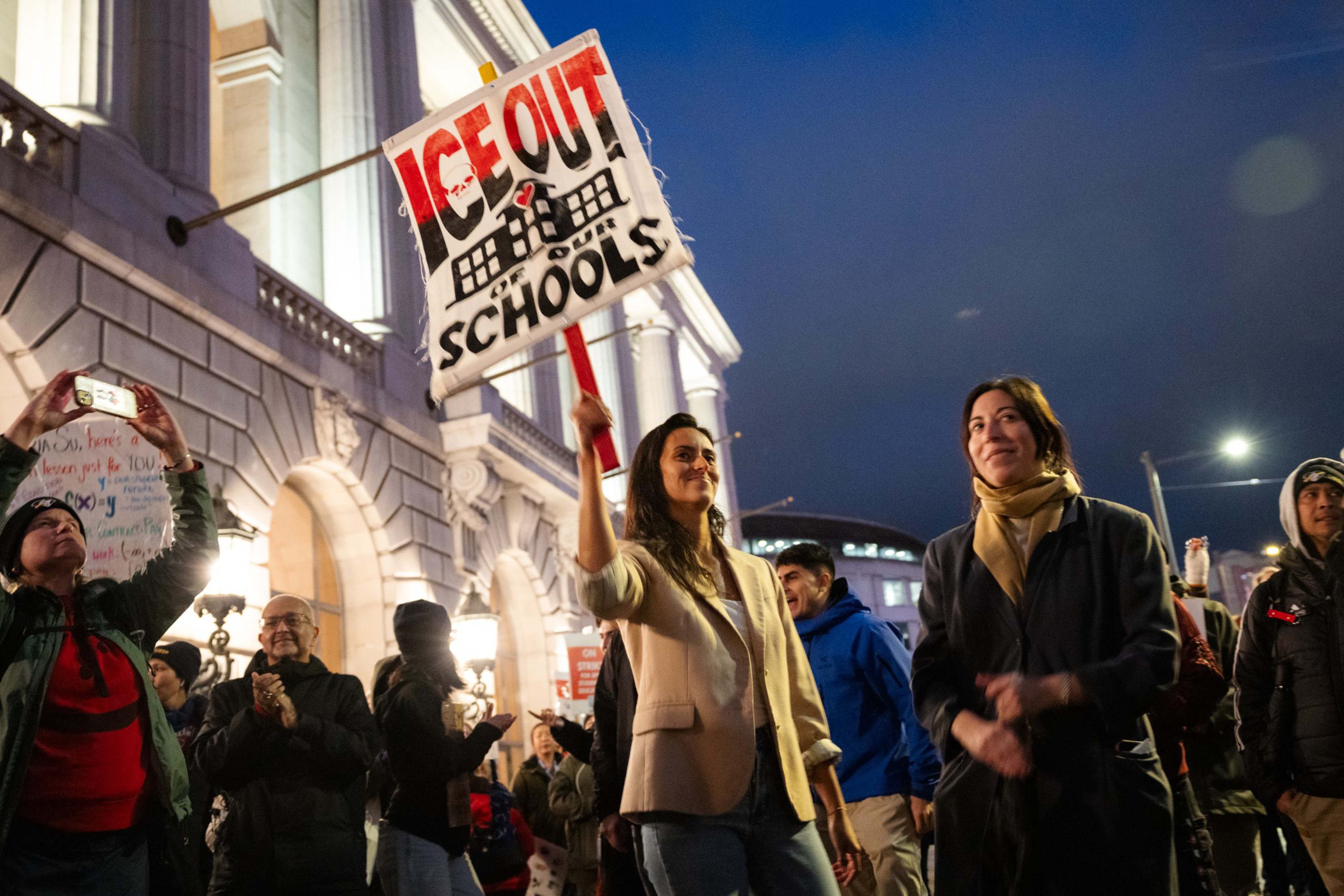 A woman holds a sign that says "ICE OUT OF SCHOOLS" while others around her smile and stand in front of a large, illuminated building at dusk.