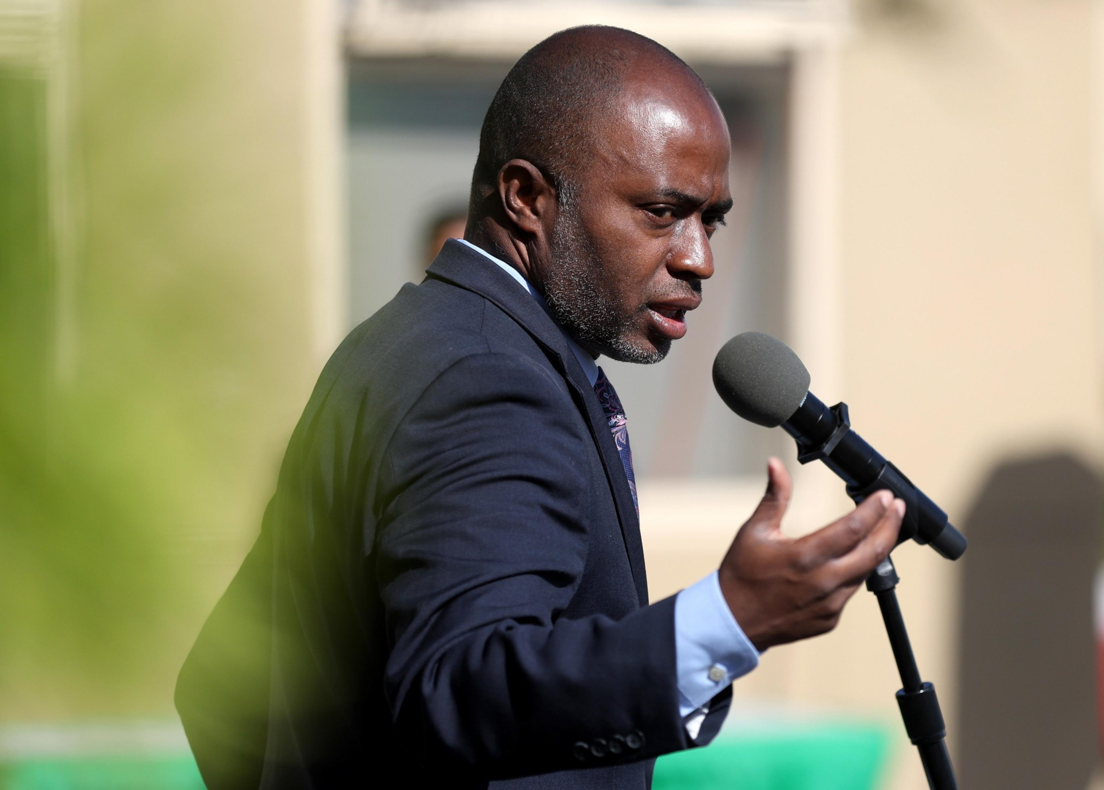 A man in a suit speaks into a microphone, gesturing with his hands, outdoors in daylight.