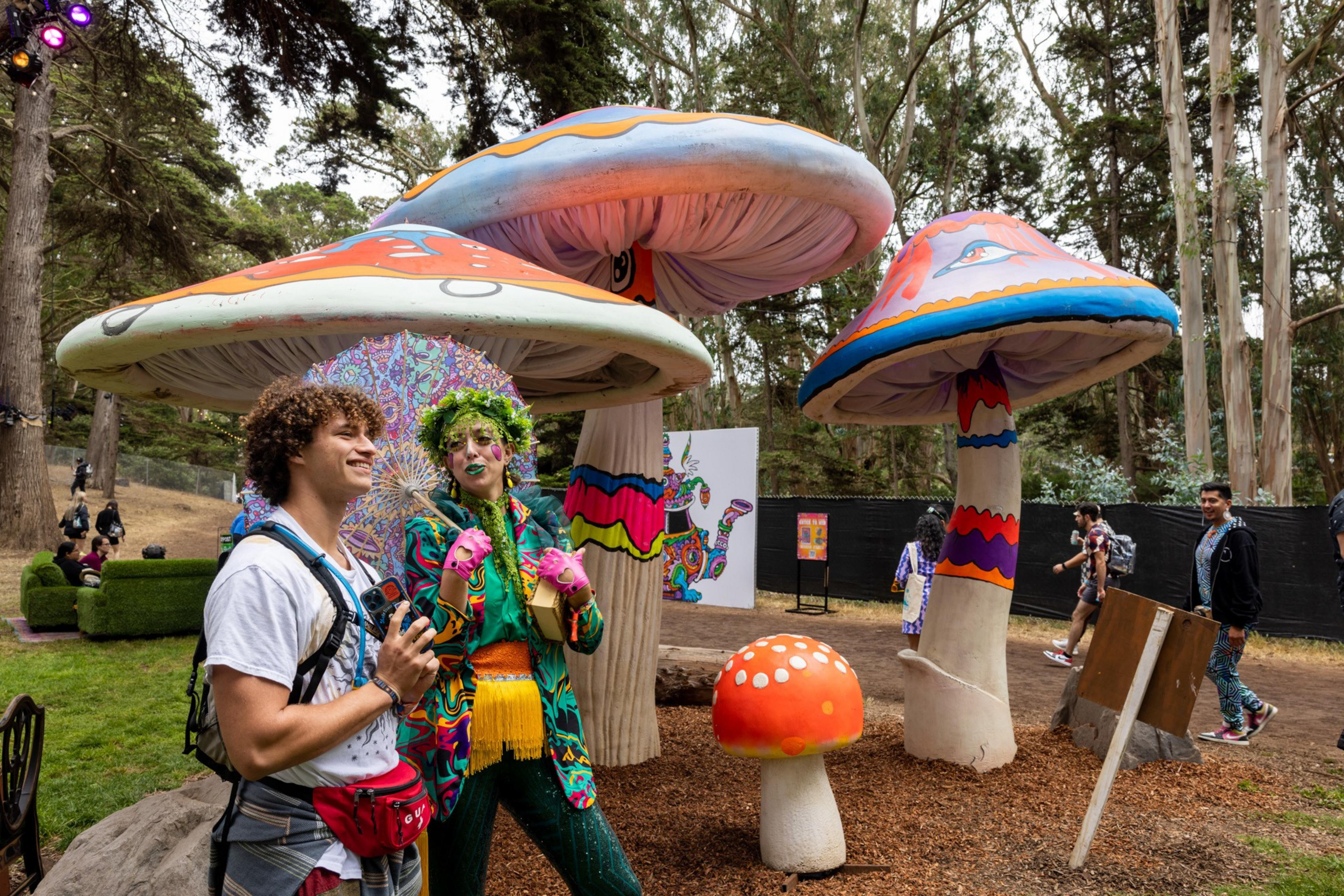 Two people in colorful costumes stand near large, whimsical mushroom sculptures in a wooded area.