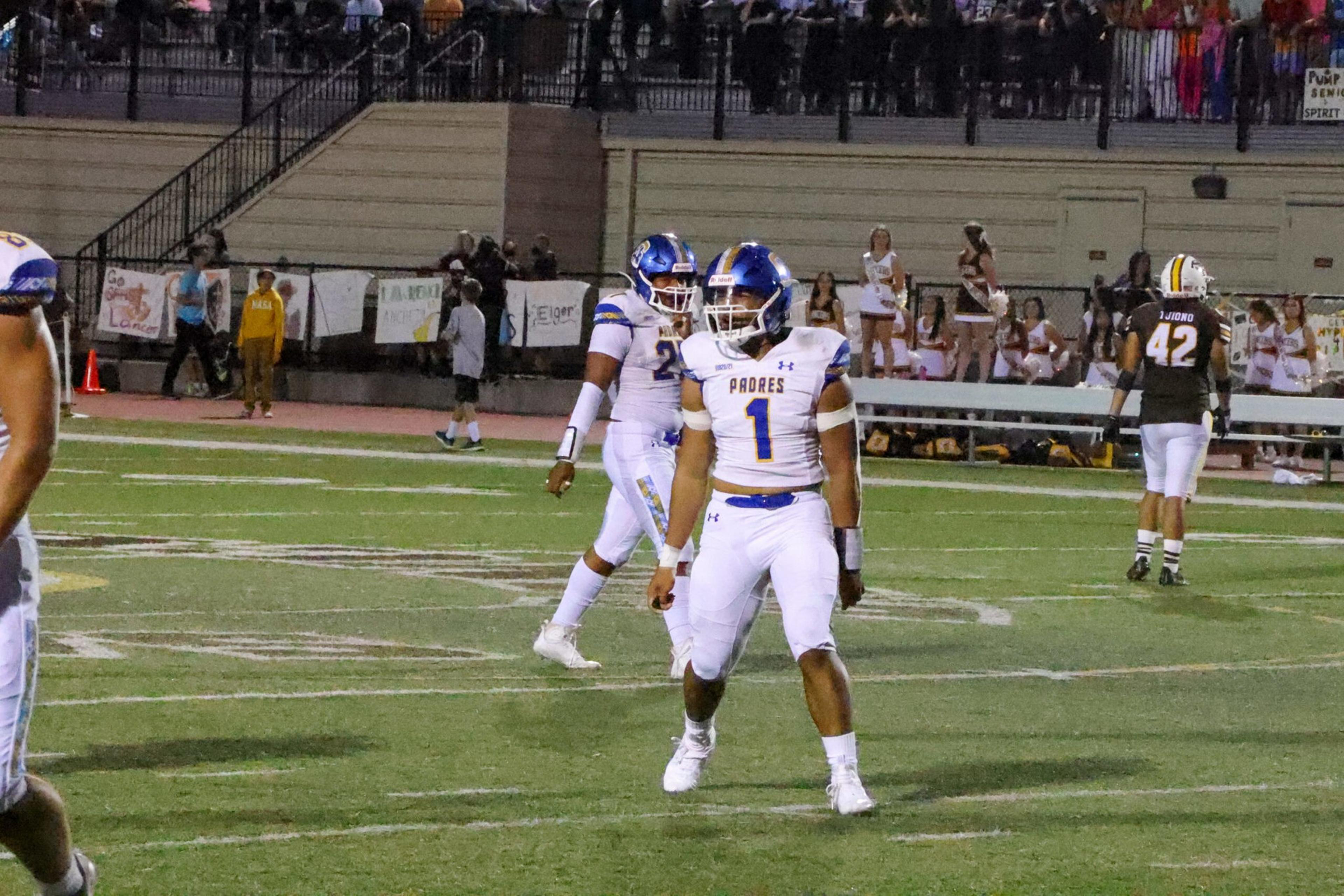 Two football players in white uniforms with blue helmets, numbered 1 and 21, stand on the field while an opposing player in black, number 42, walks away.