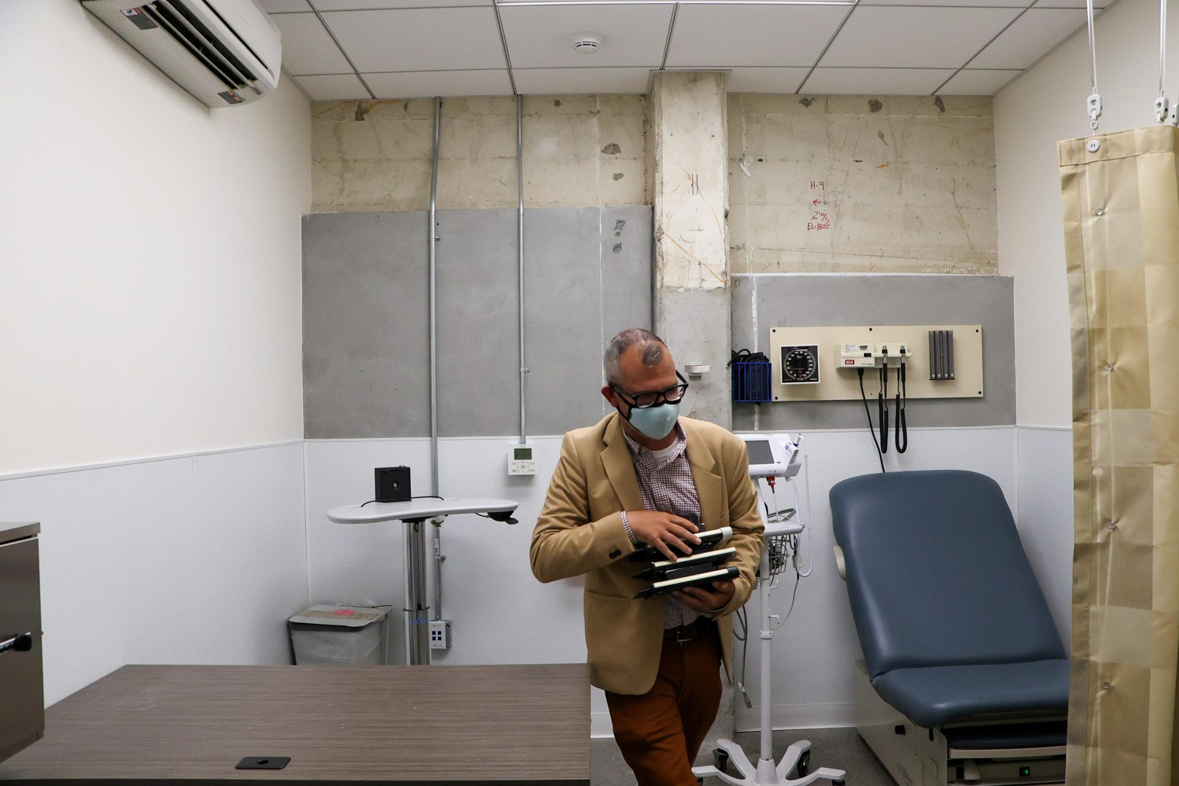 A man wearing a mask and glasses holds a stack of folders in a small, sparsely furnished medical exam room with a chair and medical equipment.