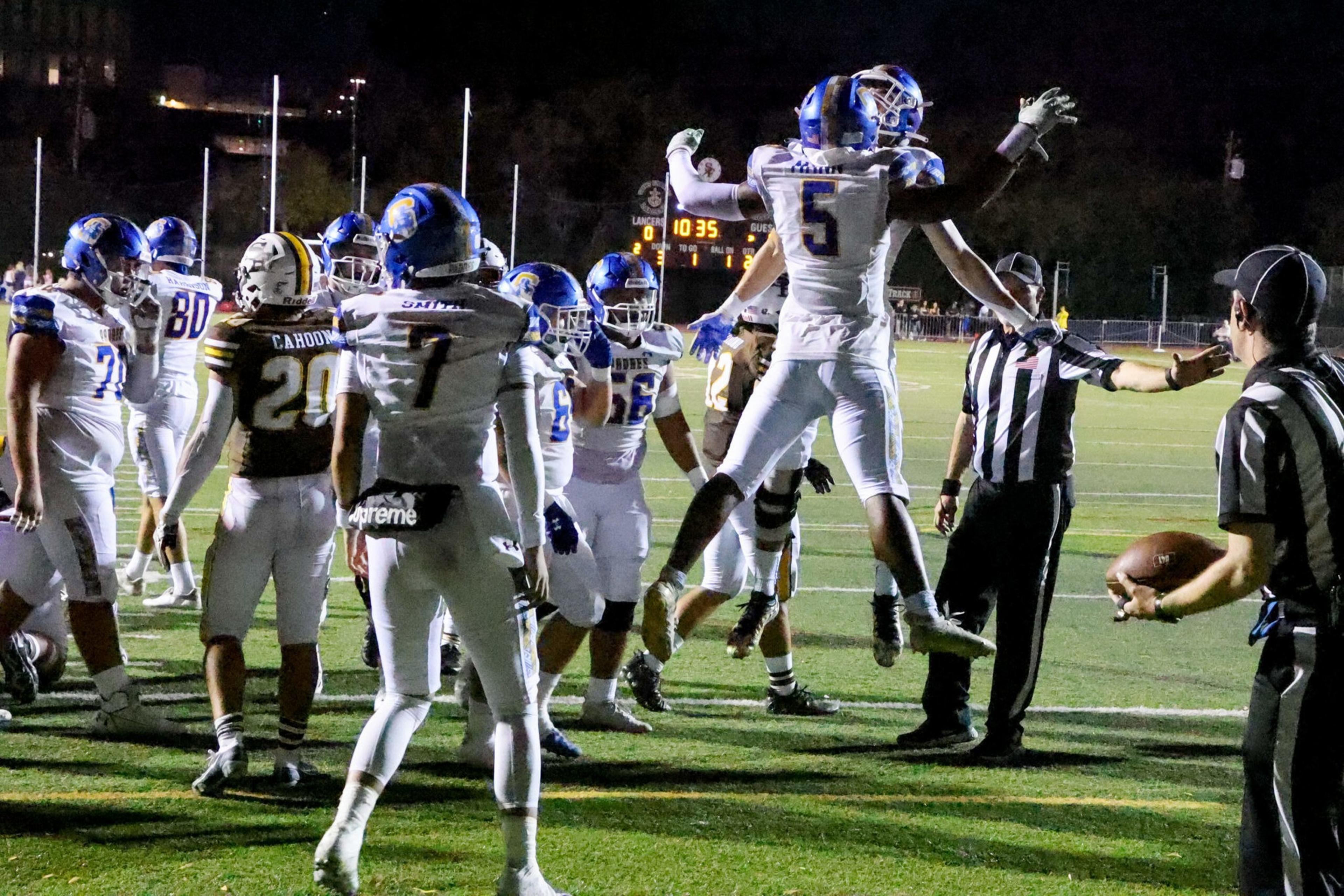 Football players in white uniforms celebrate mid-air on a field under lights, while a player in black and yellow stands nearby and referees hold the ball.